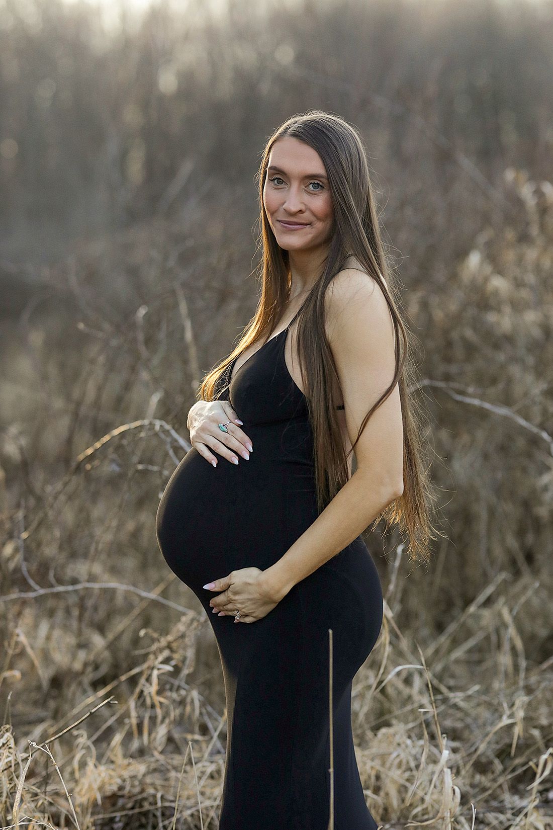 Maternity portrait session in black dress at golden hour in rustic field setting.