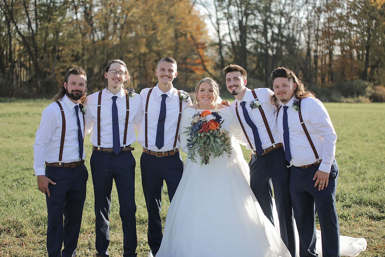 Wedding party poses together outdoors wearing navy and white attire with suspenders.