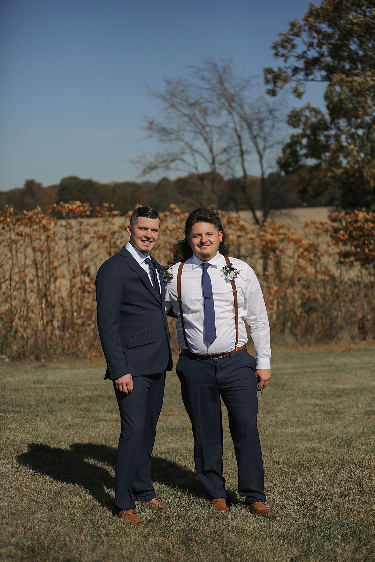 A couple in formal attire stand together in a field with fall foliage in the background.