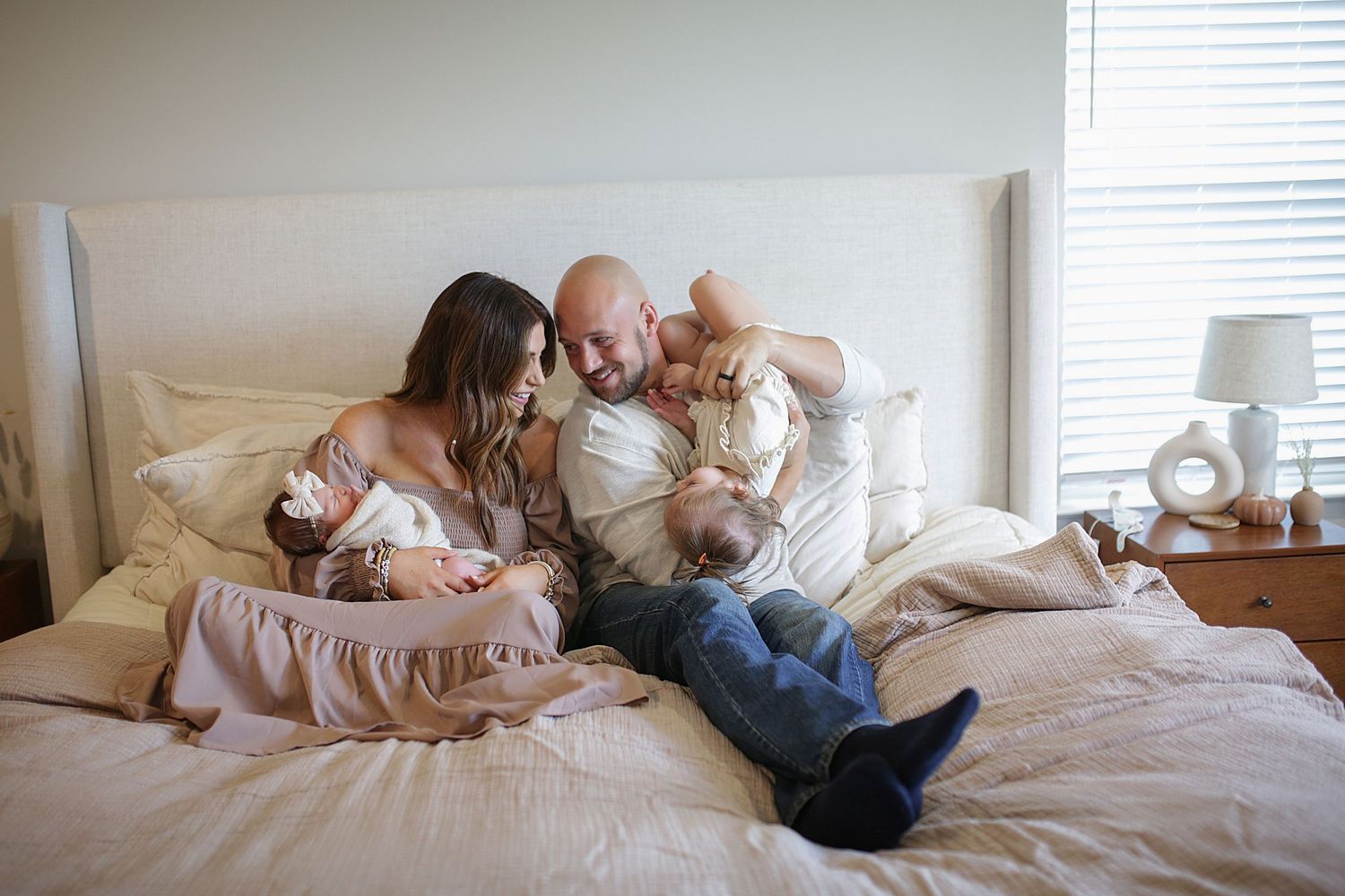A family lounges together on a white bed, sharing an intimate moment in natural lighting with muted neutral tones.