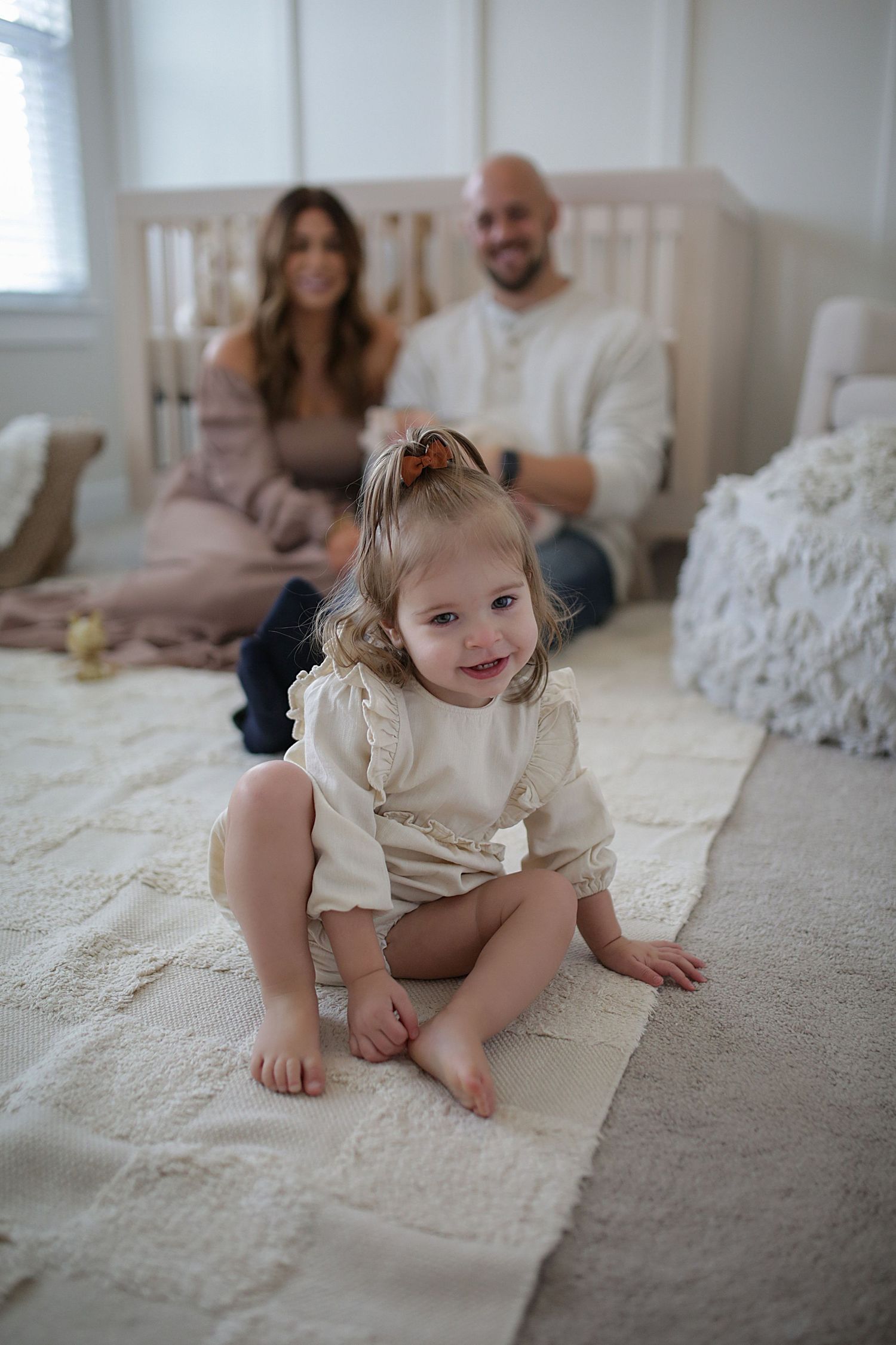Little child in cream-colored outfit sits playfully on carpet in bright bedroom with family in background.