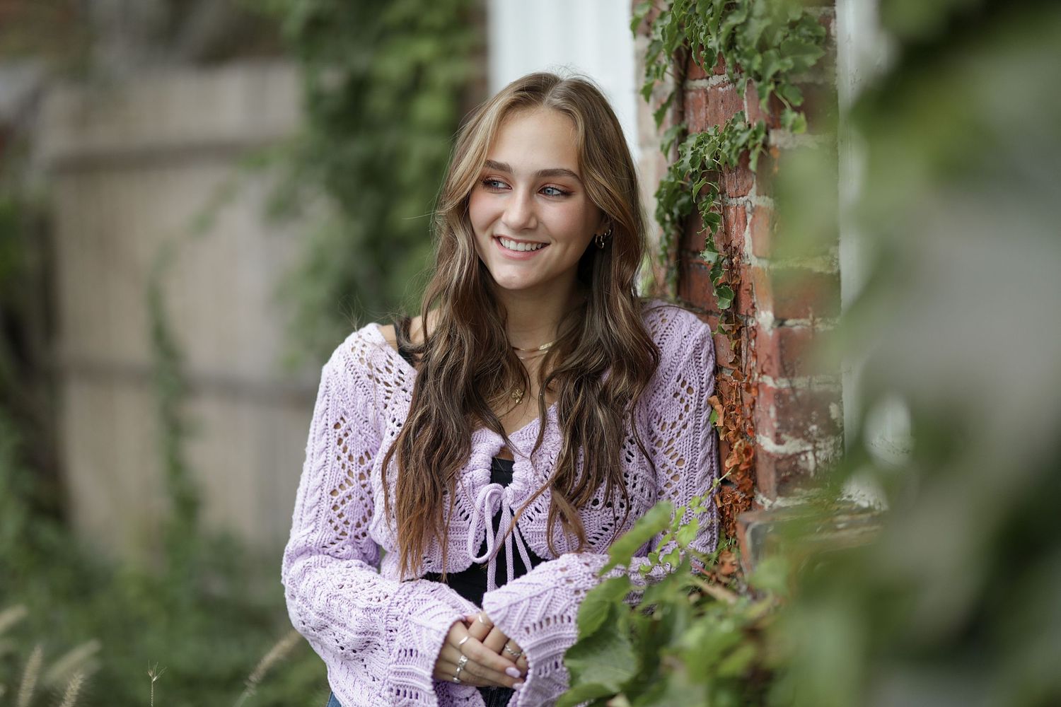 Smiling woman in lavender knit sweater poses against brick wall with greenery in garden setting.