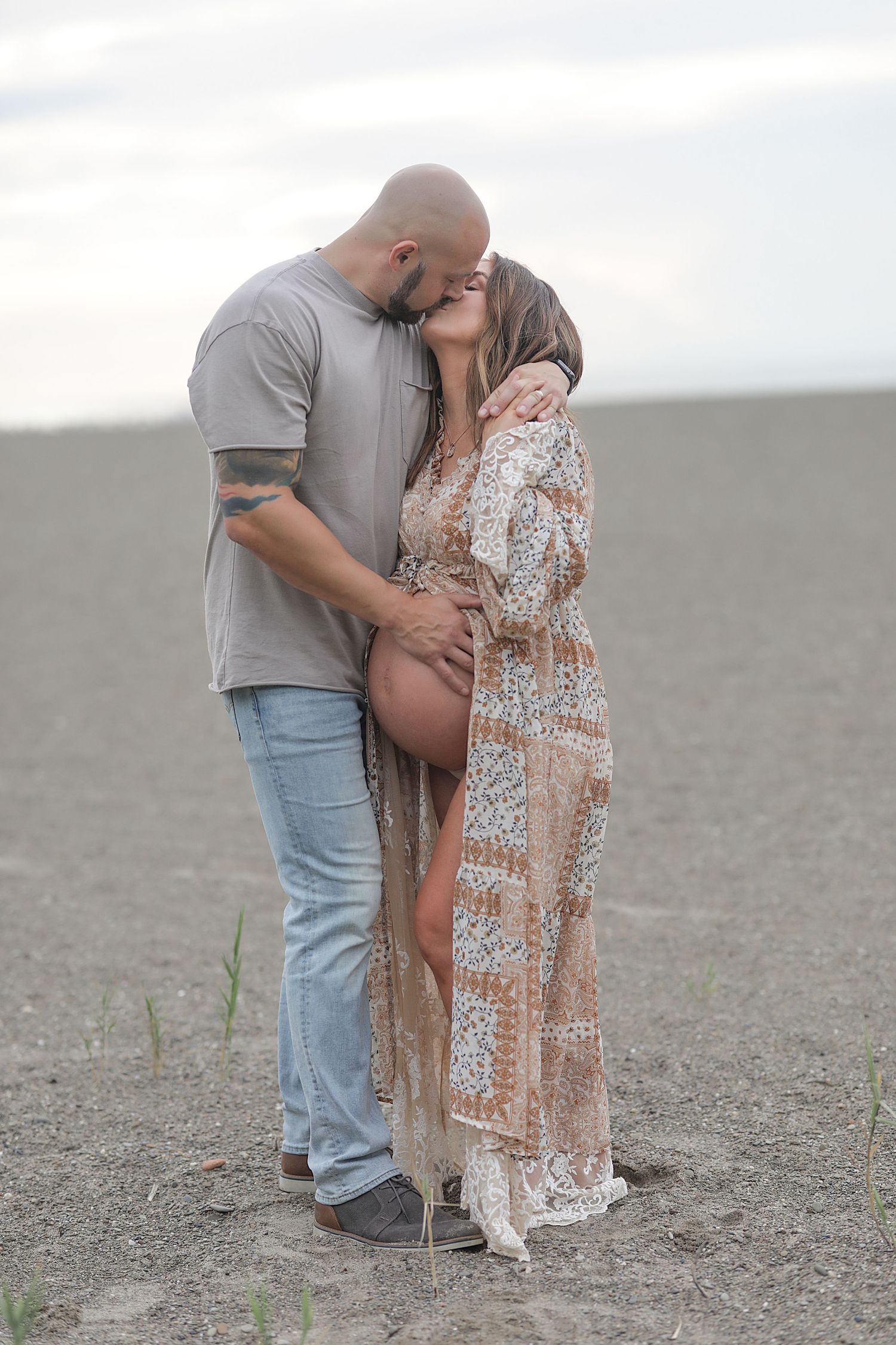 A romantic maternity photo of a couple embracing on a barren field under an overcast sky.