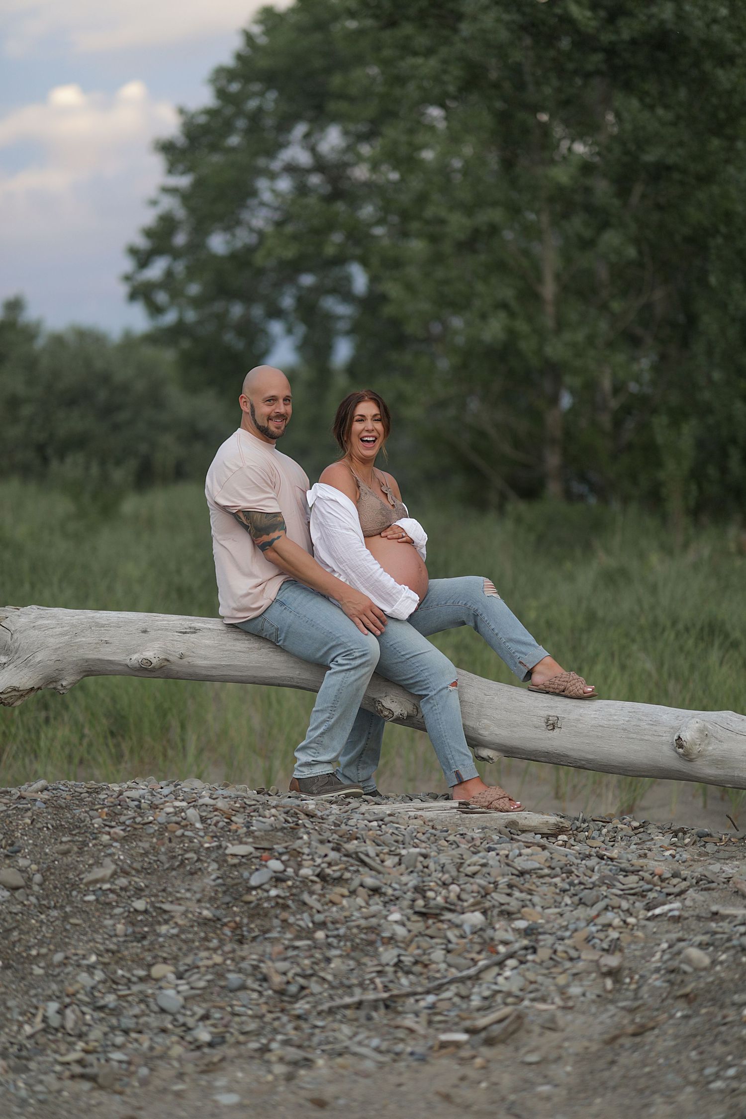 Couple sits together on fallen log during maternity photoshoot in natural outdoor setting with trees in background.