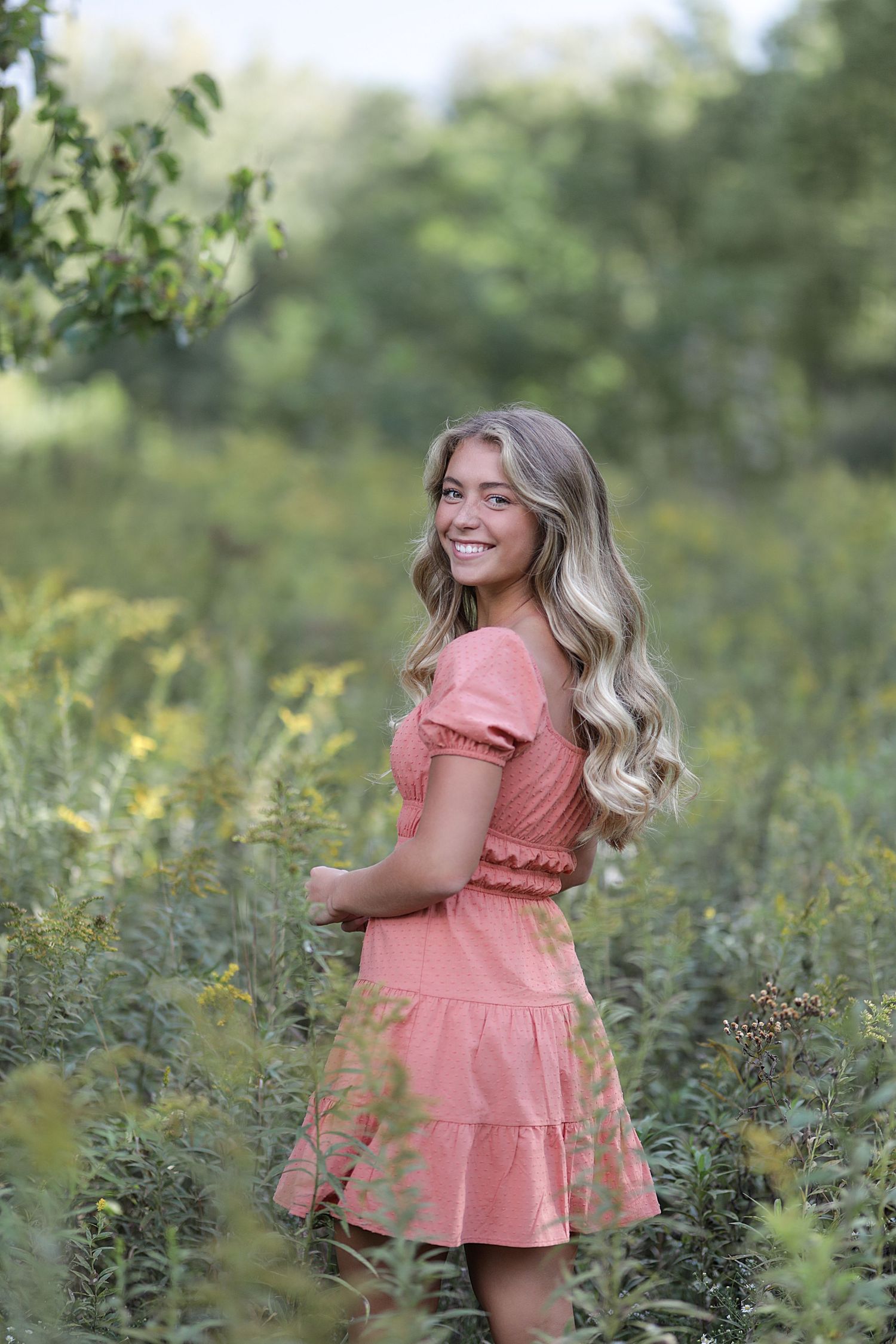 A person wearing a pink ruffled summer dress poses outdoors among wildflowers in a natural setting.