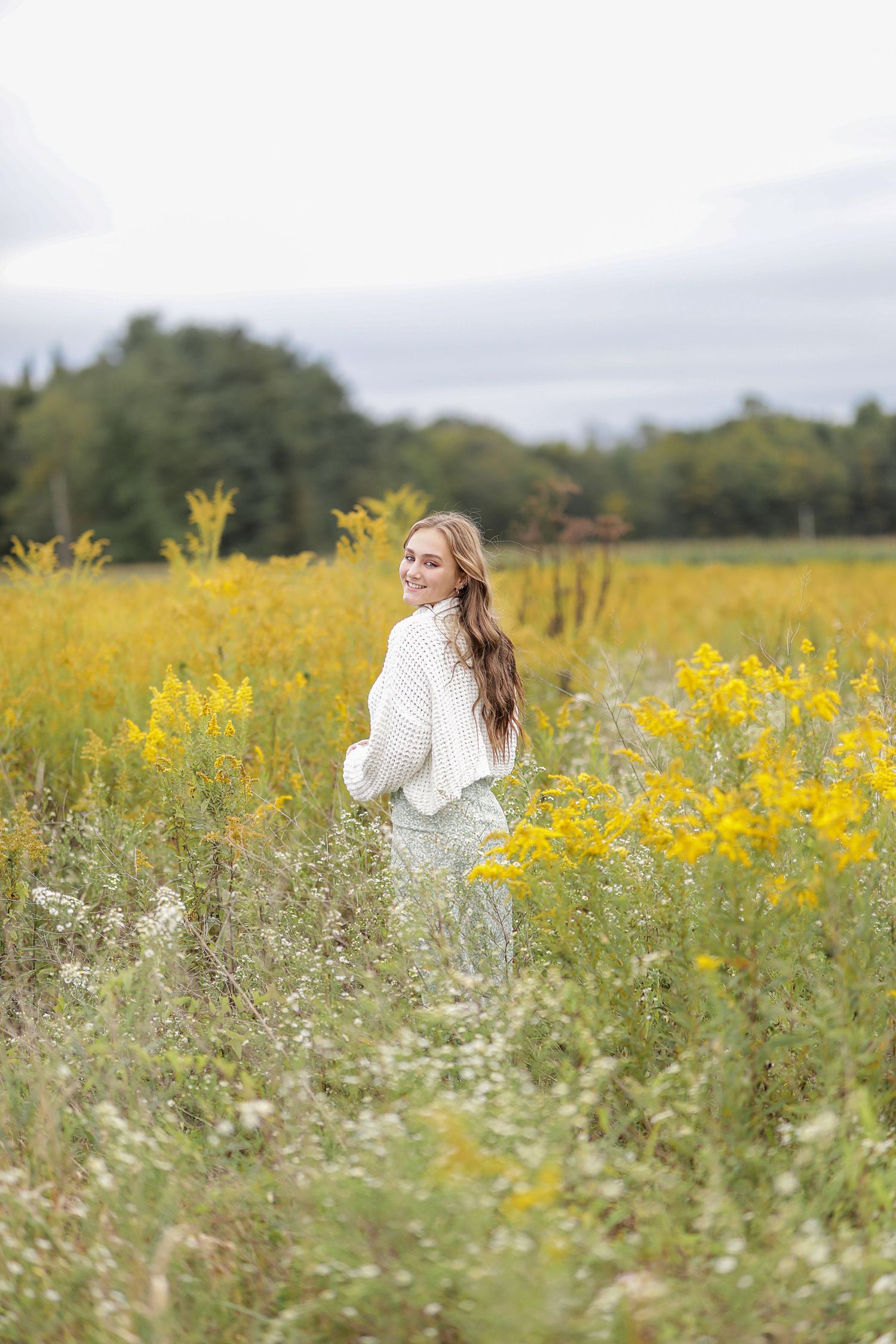 Someone in a cozy white sweater stands in a vibrant meadow of yellow wildflowers and tall grass on an overcast day.