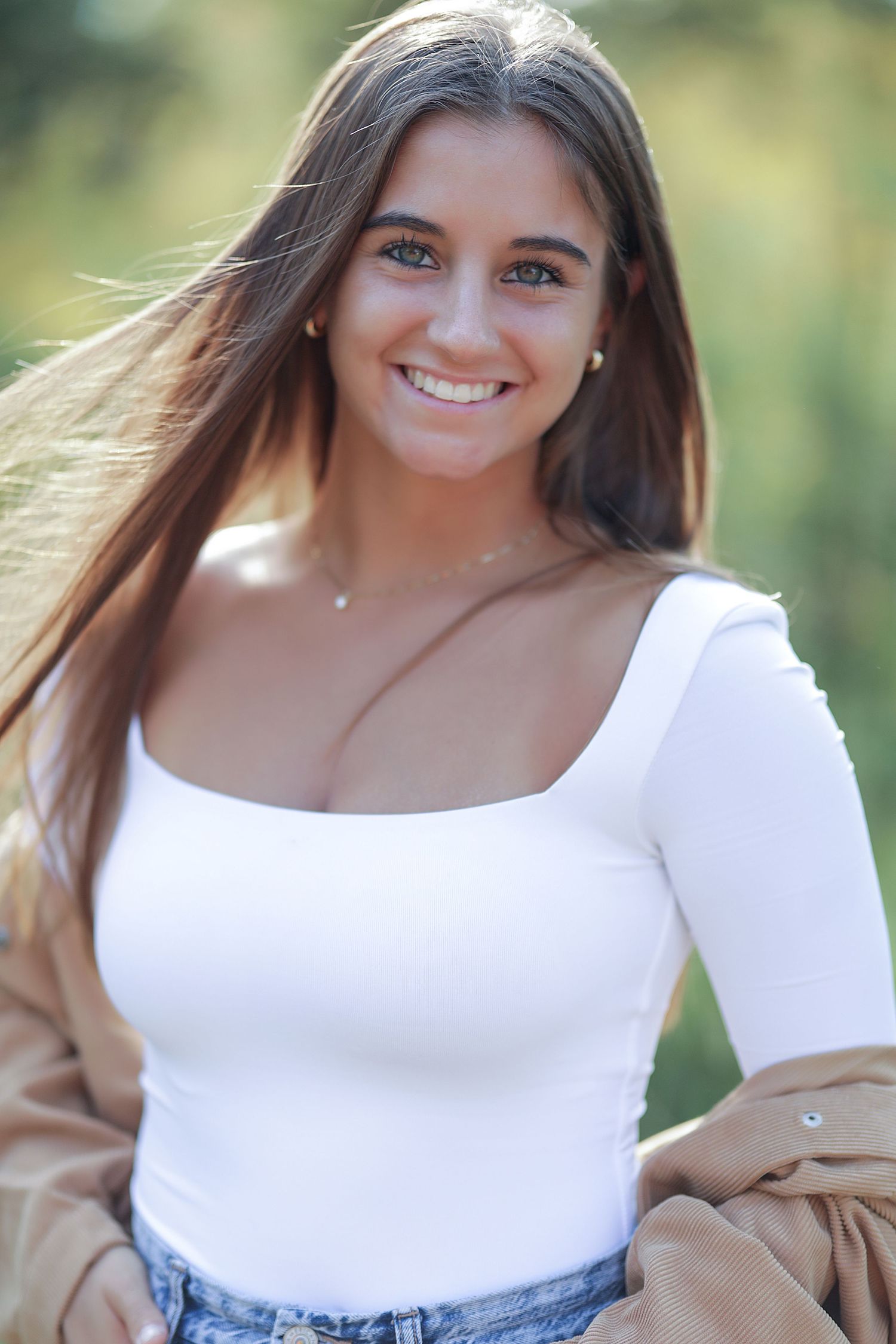 A portrait taken outdoors showing a bright smile, long brown hair, white top and tan jacket against a natural blurred background.