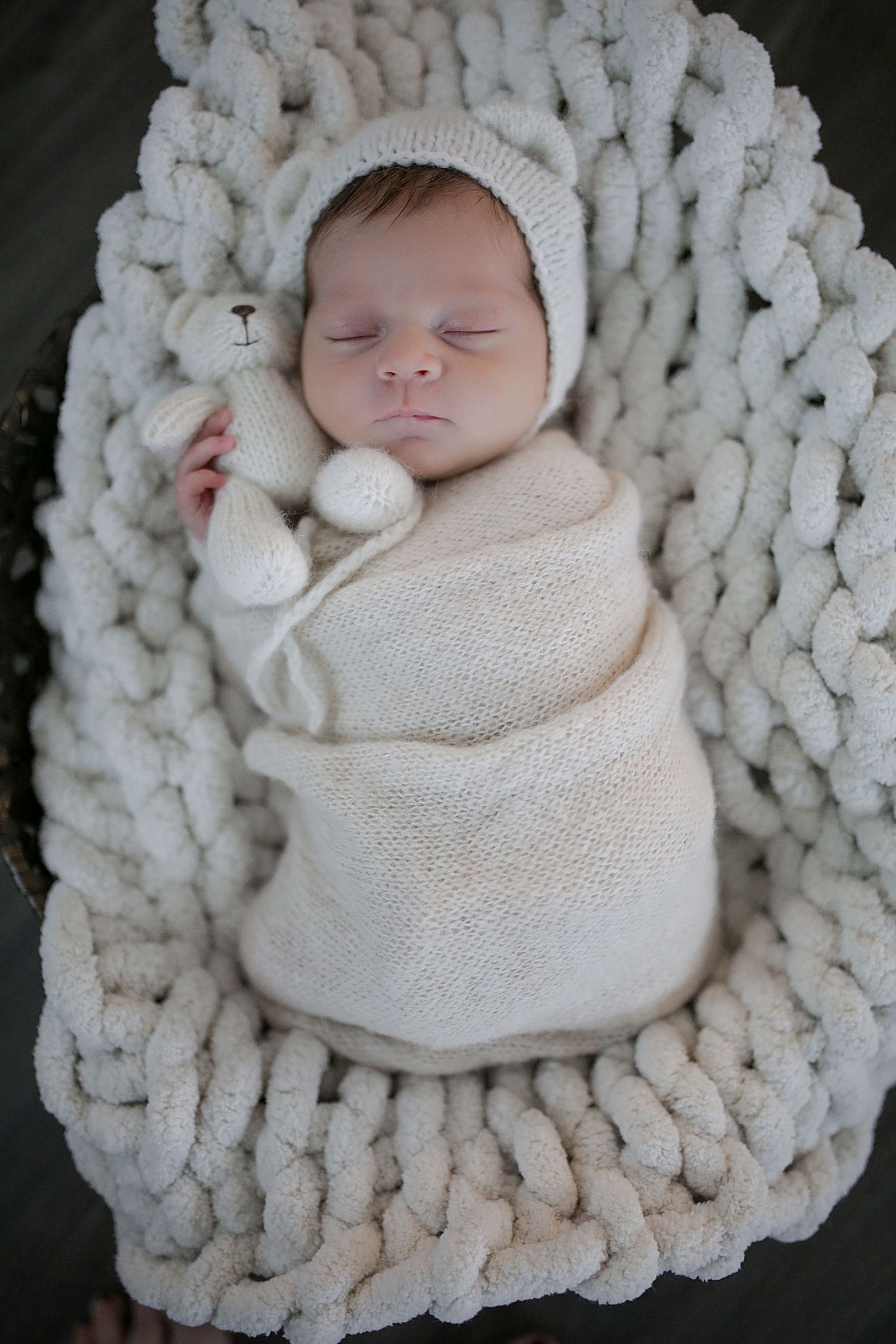 Newborn baby swaddled in white knitted wrap lying on chunky knit cream-colored blanket in artful photo series.
