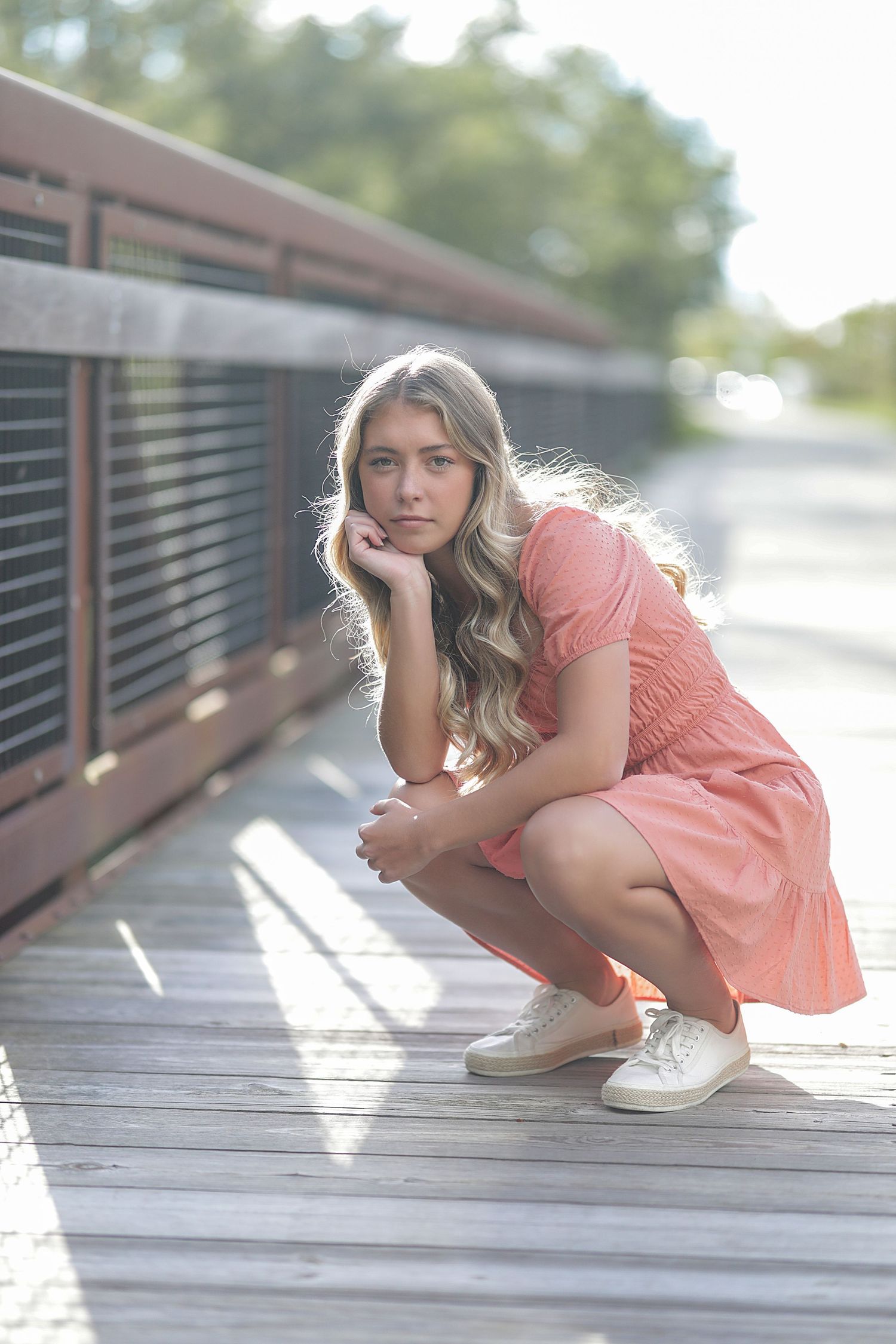 Girl in peach summer dress crouching on wooden boardwalk wearing white sneakers.