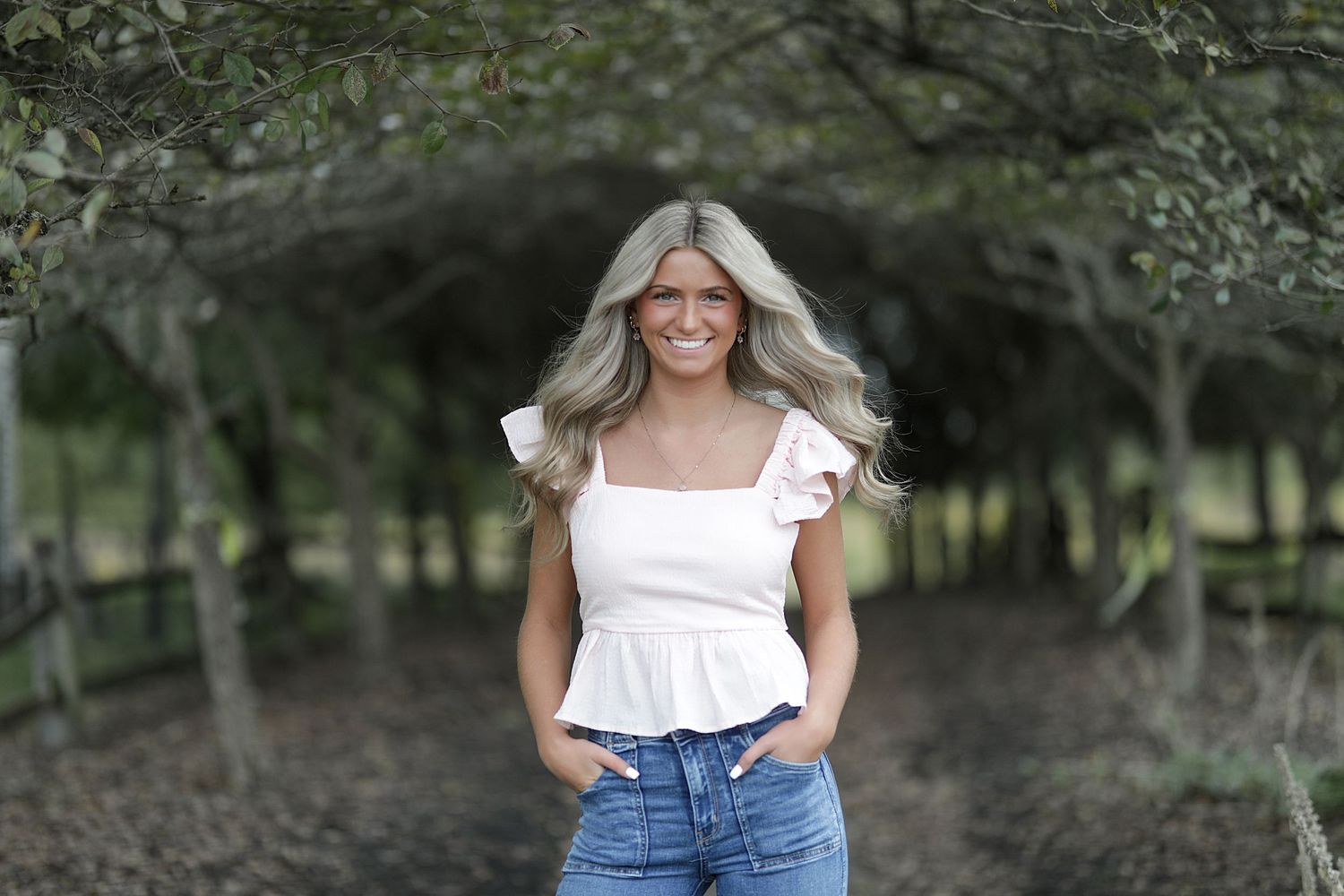 Blonde woman wearing white ruffle top and jeans stands smiling on tree-lined path.