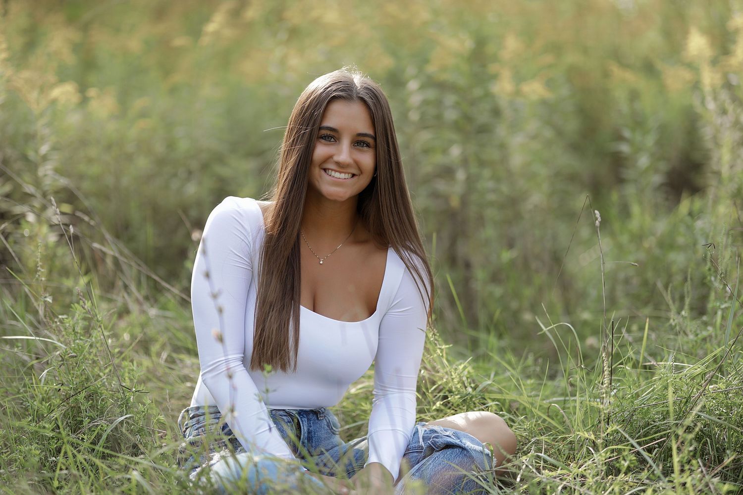 Young person in white top and jeans sitting in a grassy meadow on a sunny day.