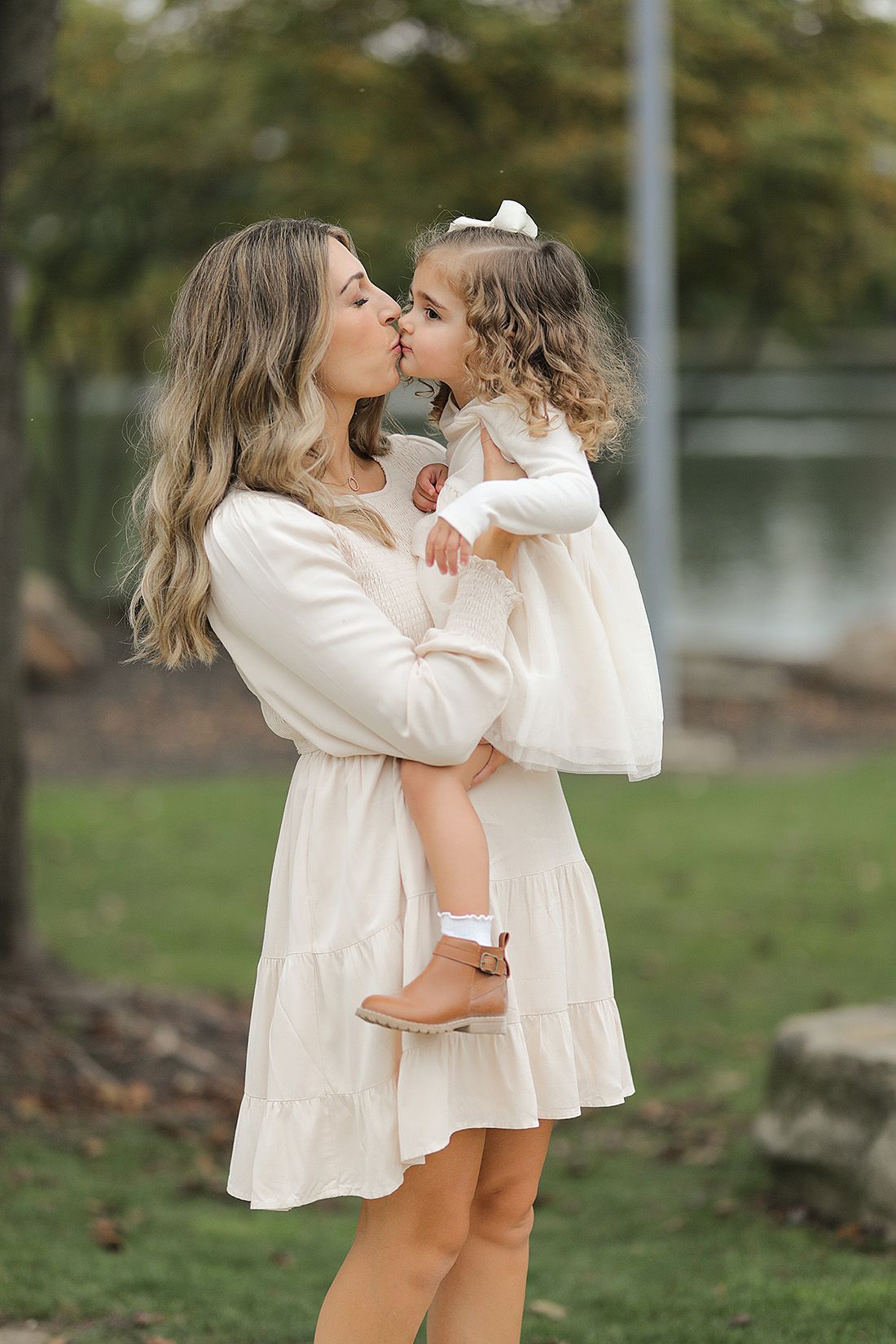Mother and daughter in coordinating cream outfits share sweet kiss during outdoor photoshoot.