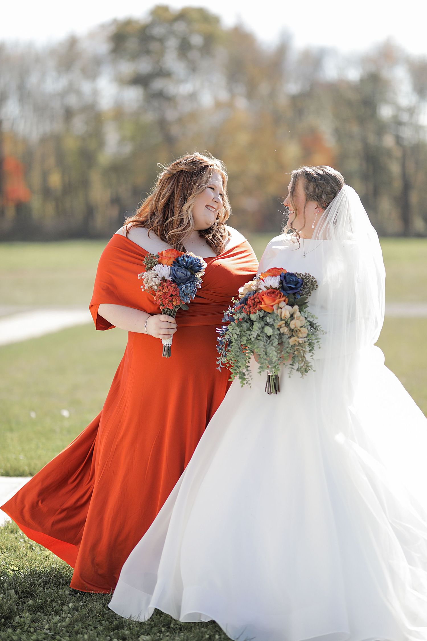 Wedding party members in bright orange dresses embrace the bride outdoors in autumn.