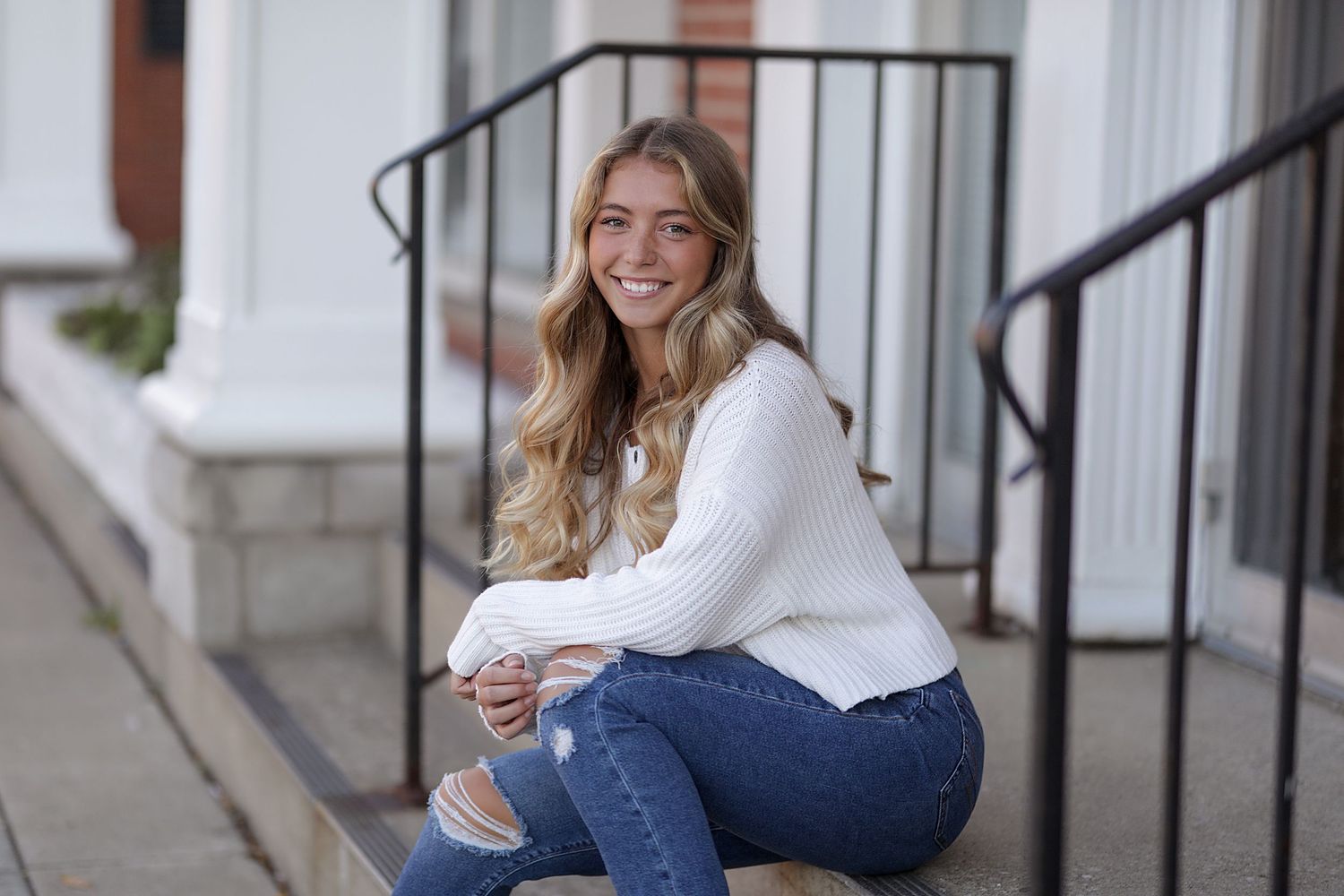 Young woman in white sweater and jeans sitting on steps while smiling at camera.