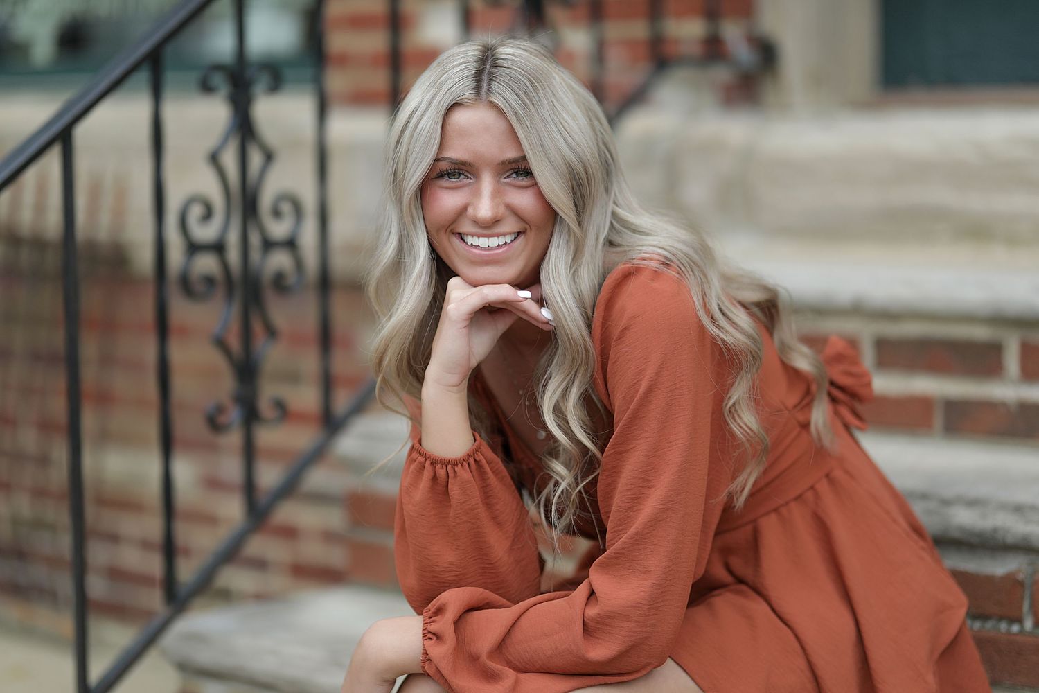 Someone with long wavy platinum blonde hair wearing a rust-colored sweater sits on outdoor steps smiling.