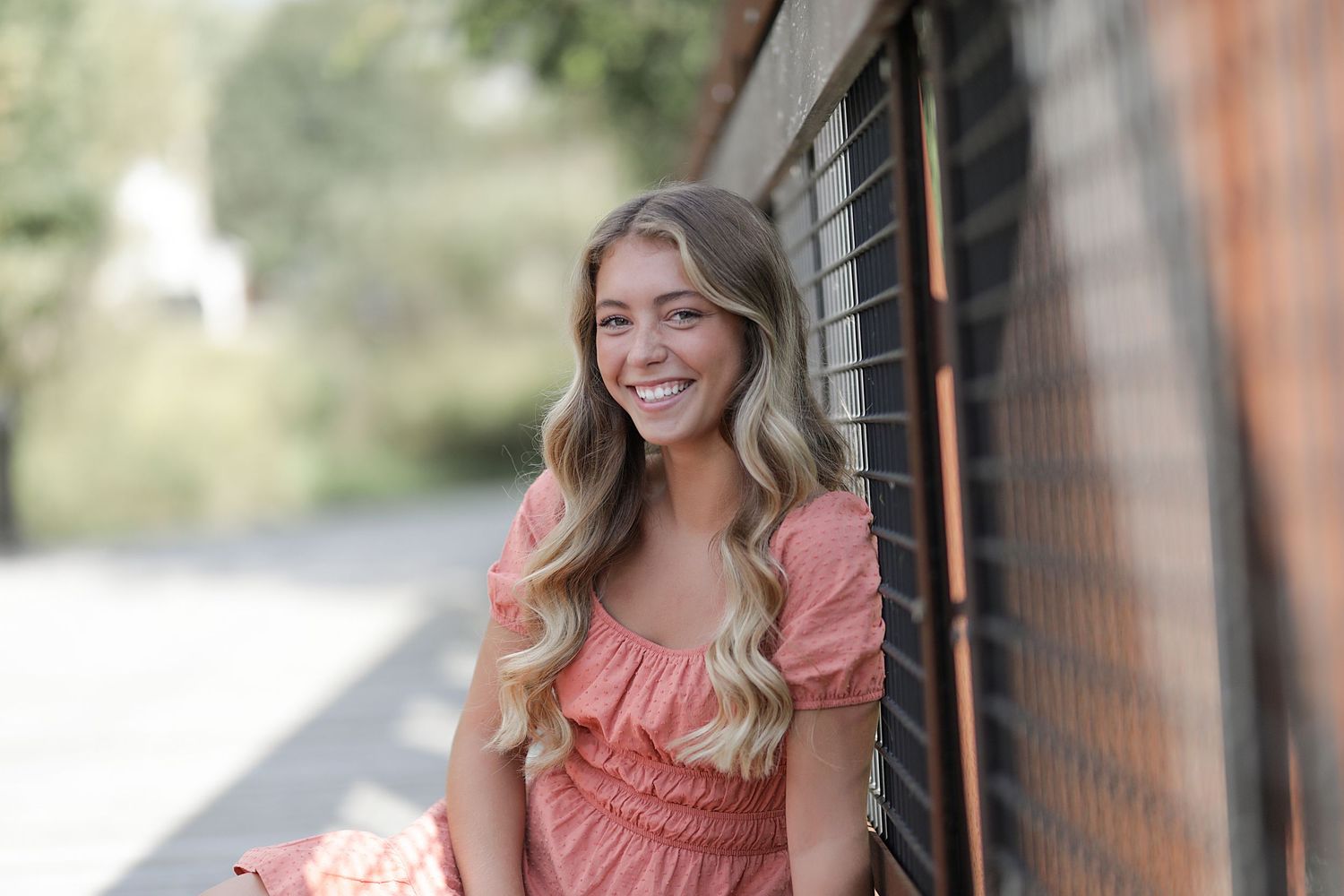 A person with long wavy blonde hair smiles while wearing a coral pink dress, sitting outdoors near a wall.