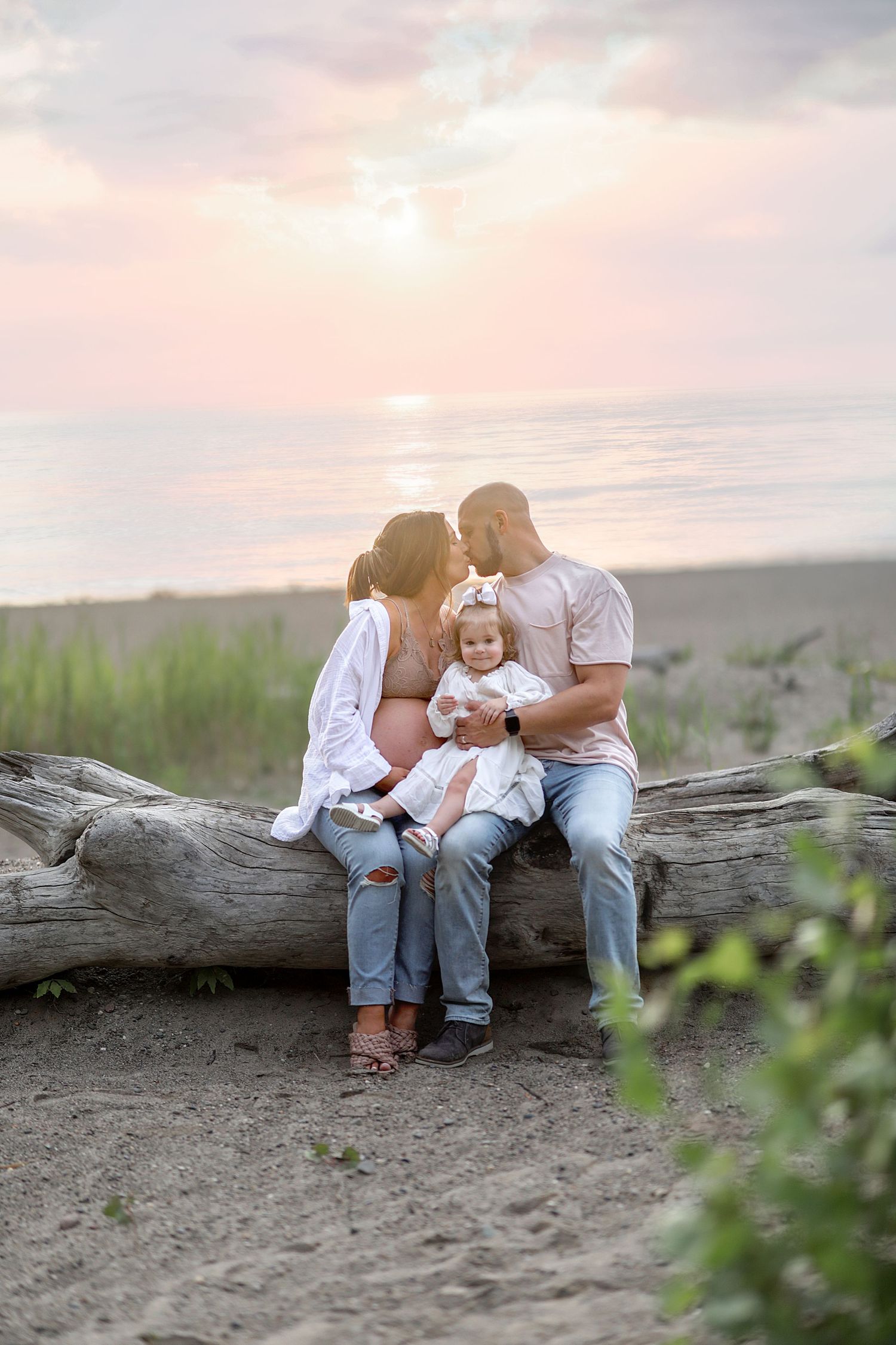 A loving family sits together on a weathered log at the beach during golden hour sunset.