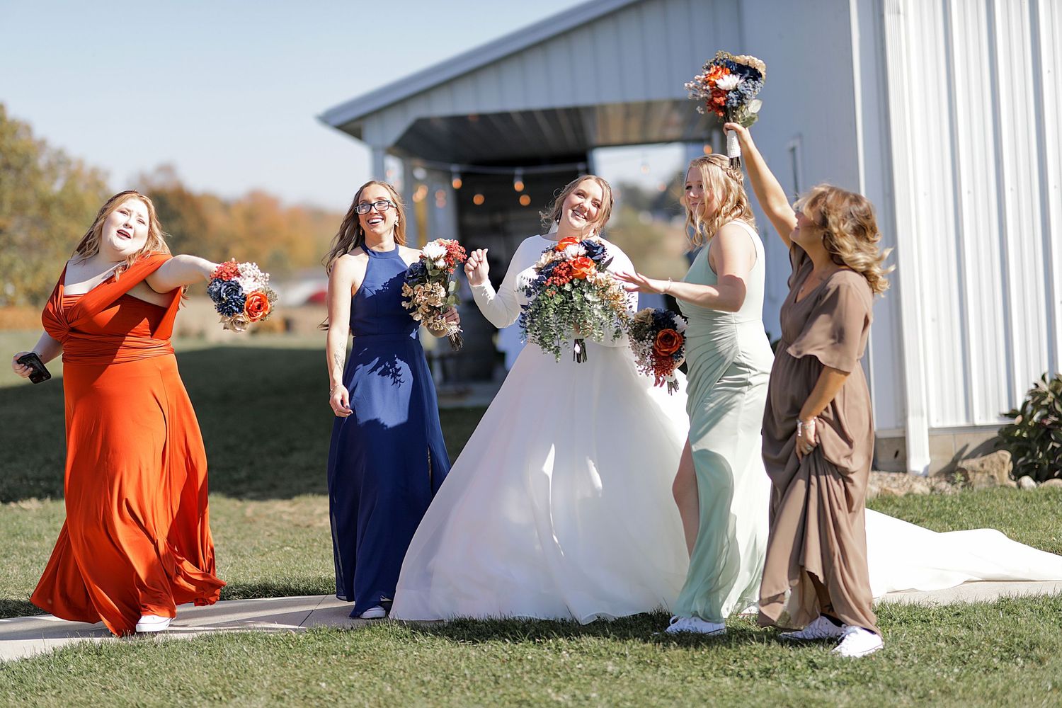 Wedding party in orange and neutral dresses gather outside a white barn.