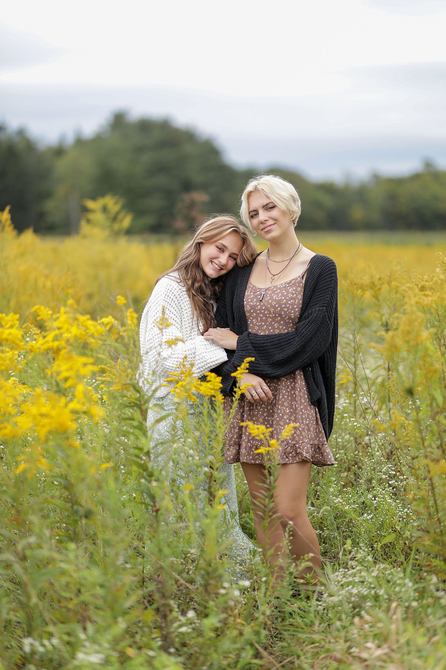 Two women in casual dresses stand together in a field of blooming yellow wildflowers on a cloudy summer day.