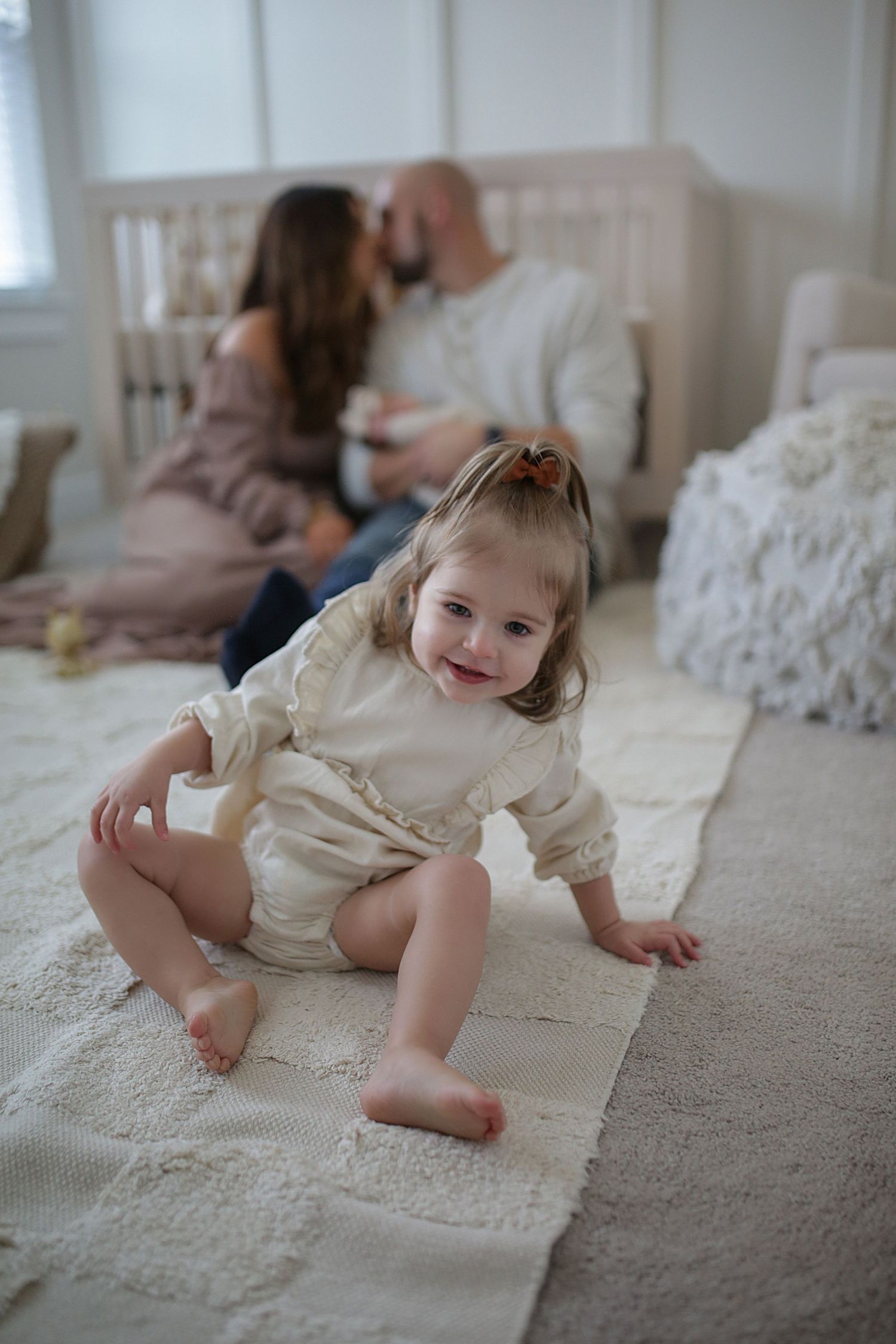A young child in a cream-colored romper sits playfully on a carpet while two people relax in the background.