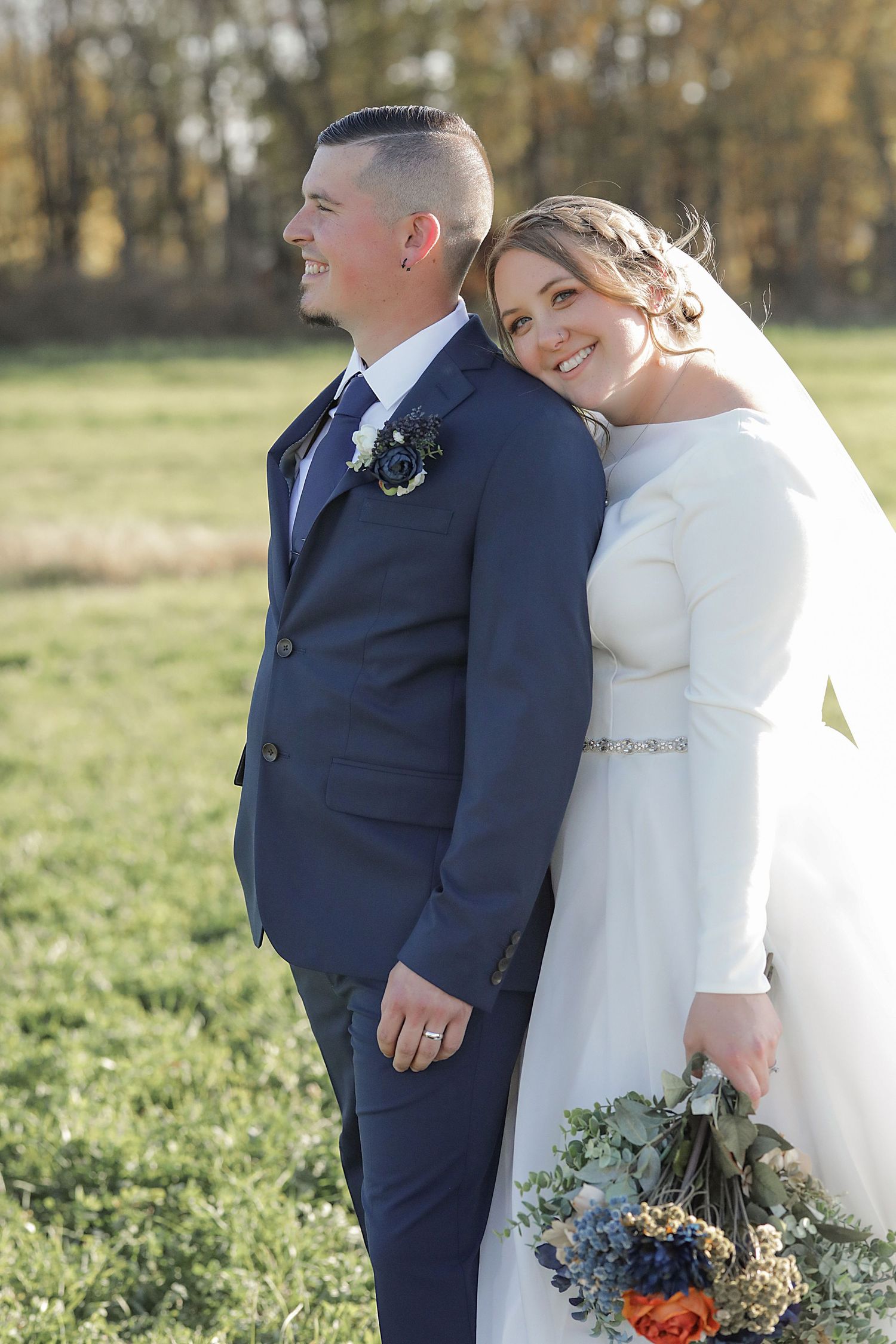 Outdoor wedding portrait of a couple in navy suit and white gown with floral bouquet against autumn trees.
