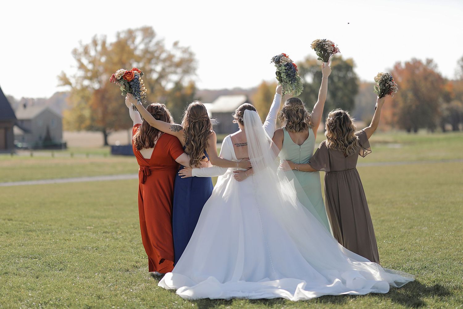 Wedding party members interact playfully on grassy field outside white barn.