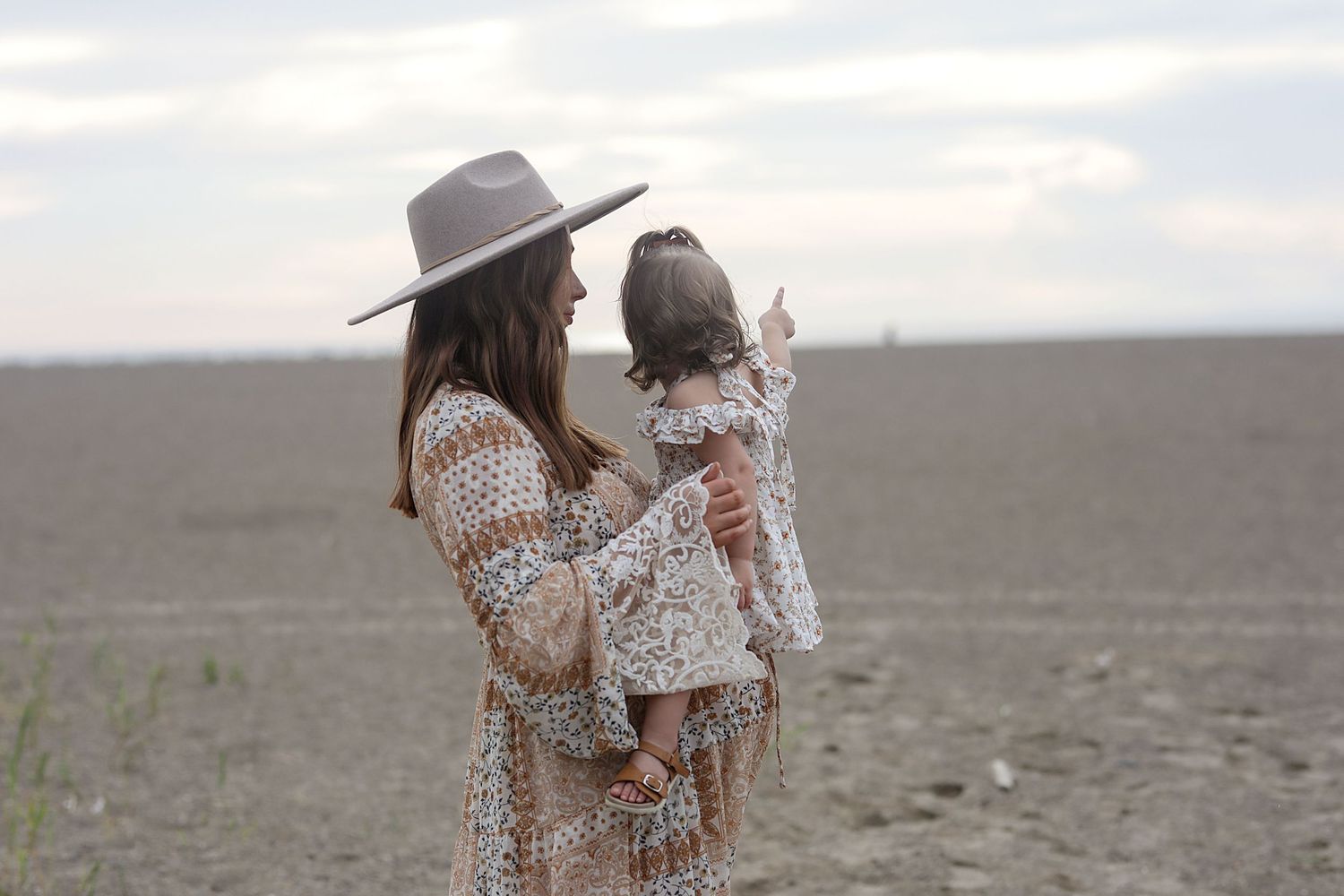 A person in a wide-brimmed hat and floral dress holds a child while standing in an open field during golden hour.