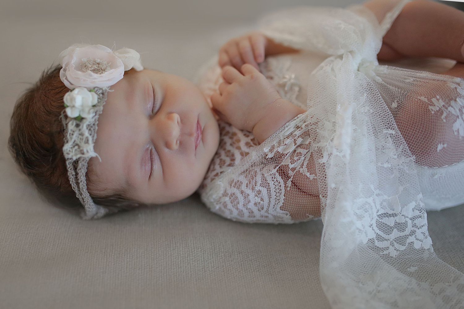 A sleeping baby wearing a white lace dress and delicate headband rests peacefully on a light surface.
