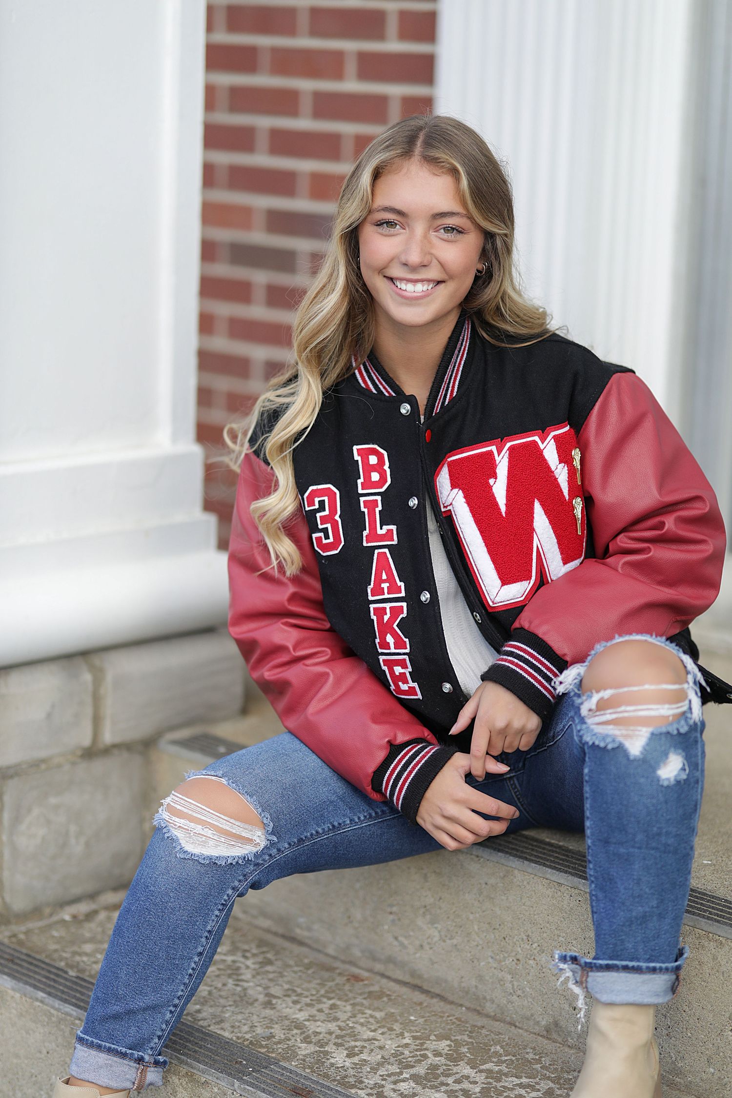 Student wearing a red and black varsity letterman jacket and distressed jeans sits on concrete steps by a brick wall.