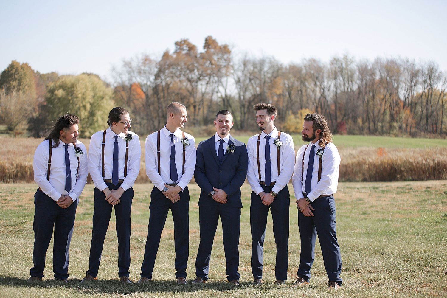A group of groomsmen in matching suspenders and ties pose with the groom in a field.