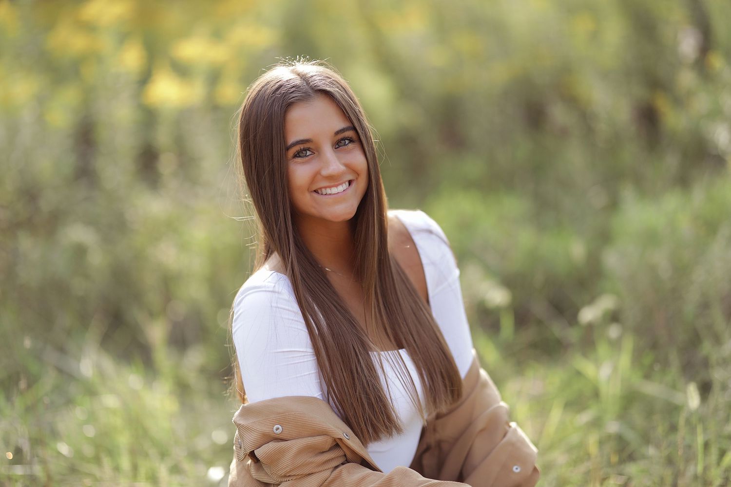 A woman with long brown hair sits outdoors smiling warmly while wearing a white shirt and tan cardigan.