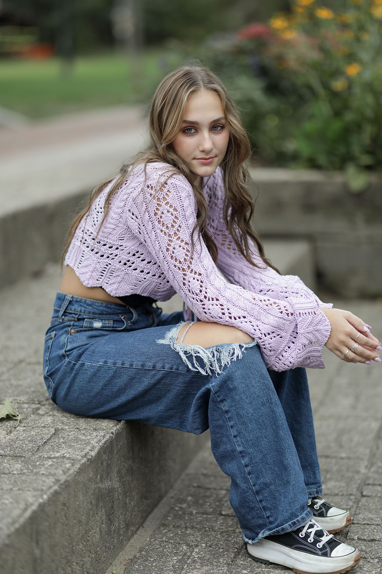 A person sits casually on concrete steps wearing a lavender knit sweater, distressed jeans, and black sneakers.