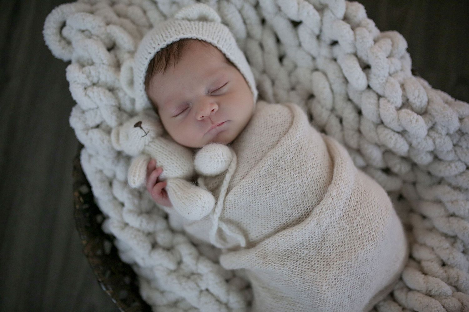 Sleeping newborn swaddled in cream-colored wrap rests peacefully on chunky knit blanket.
