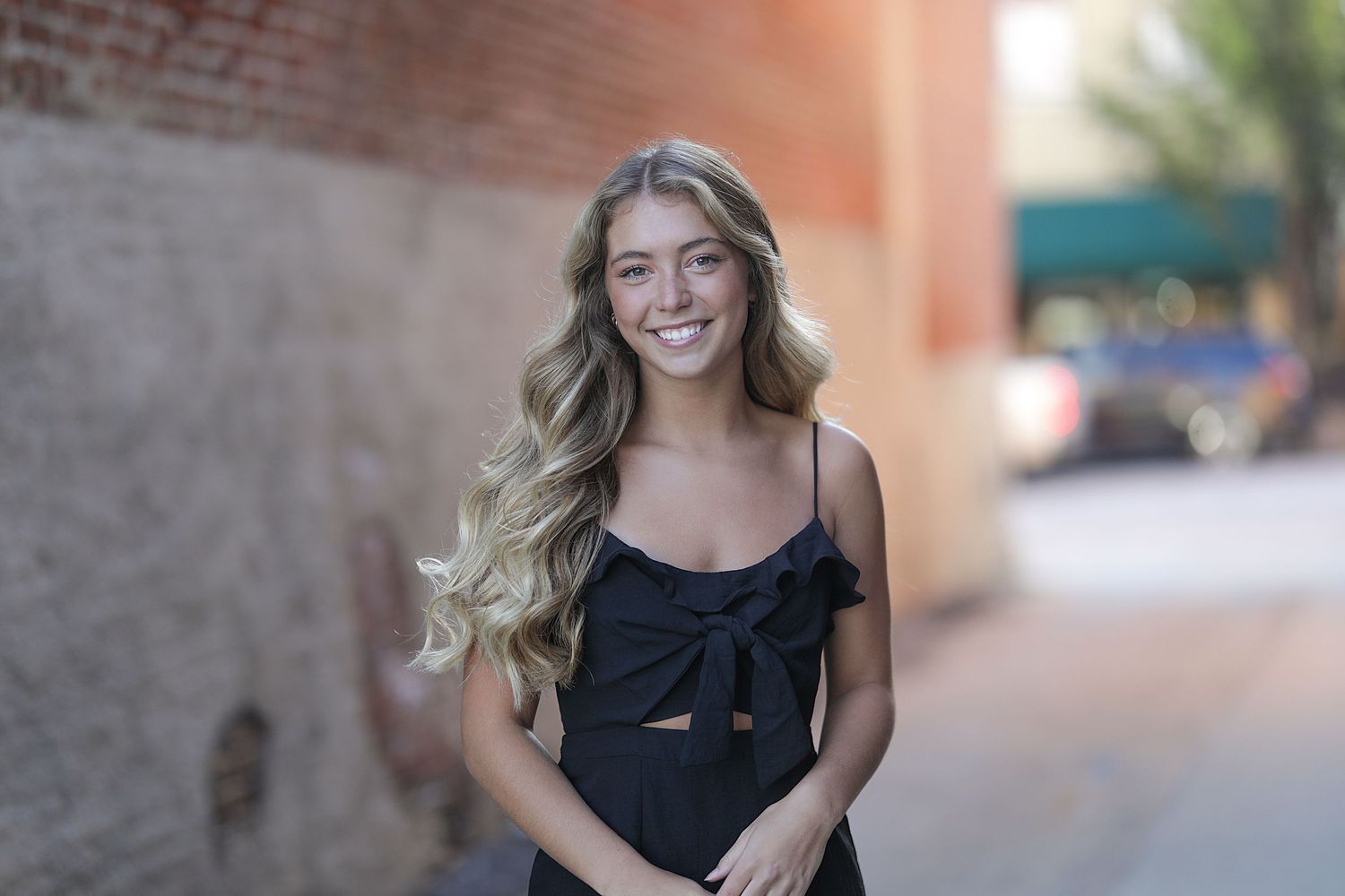 A person in a black top stands outdoors against a blurred brick wall with long blonde wavy hair.