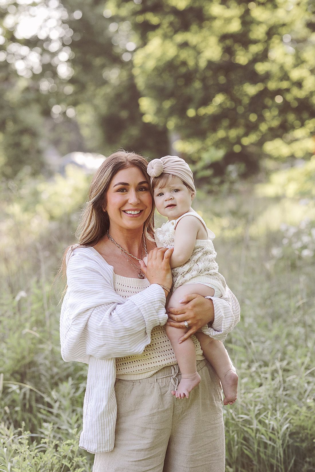 Mother in white sweater holding baby in sunny outdoor setting with natural bokeh background.