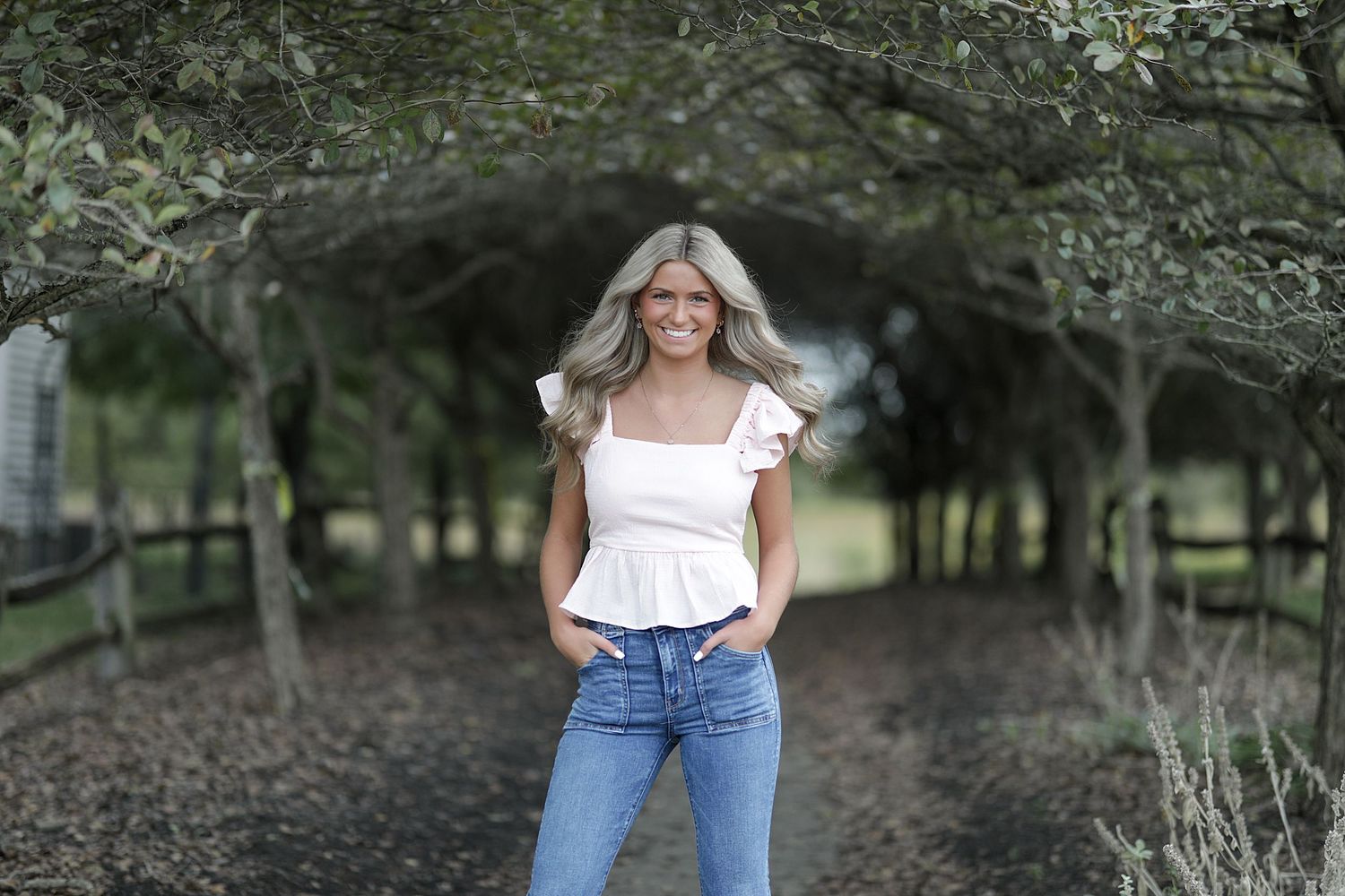 A casual outdoor portrait featuring a white ruffled top paired with classic blue jeans under a tree-covered path.