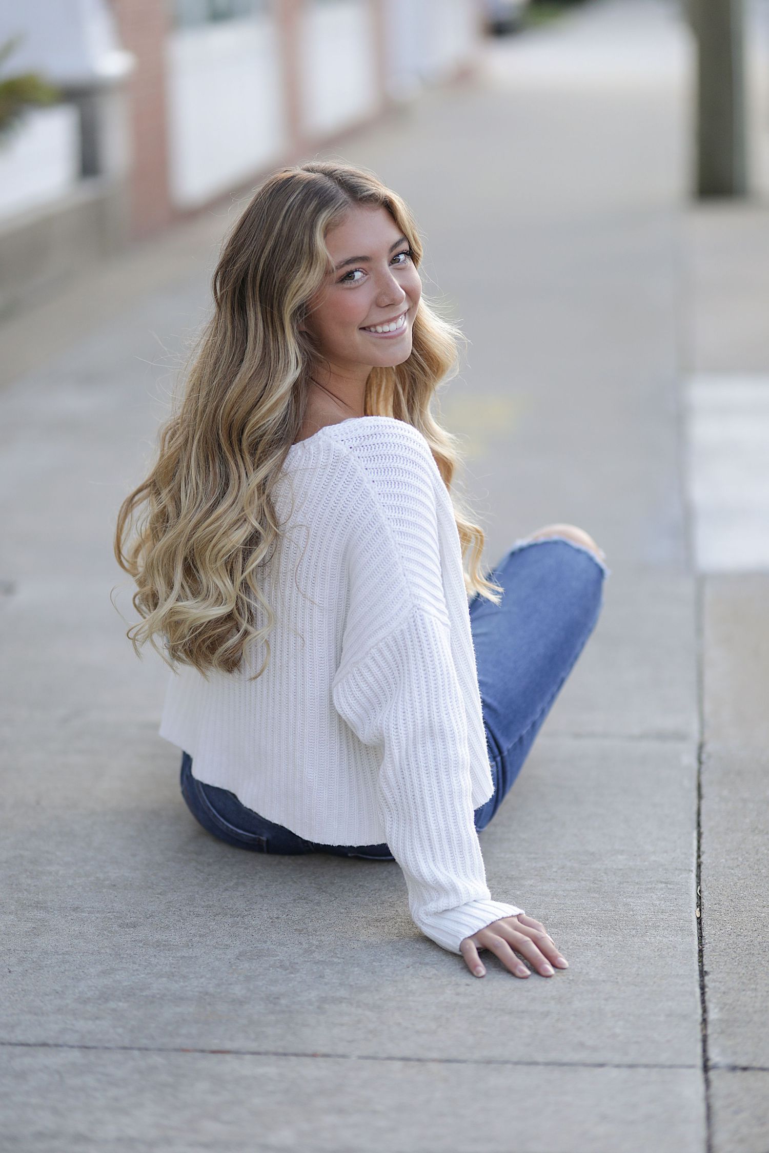 Casual portrait of someone in a white sweater and jeans sitting on concrete while smiling and looking over shoulder.