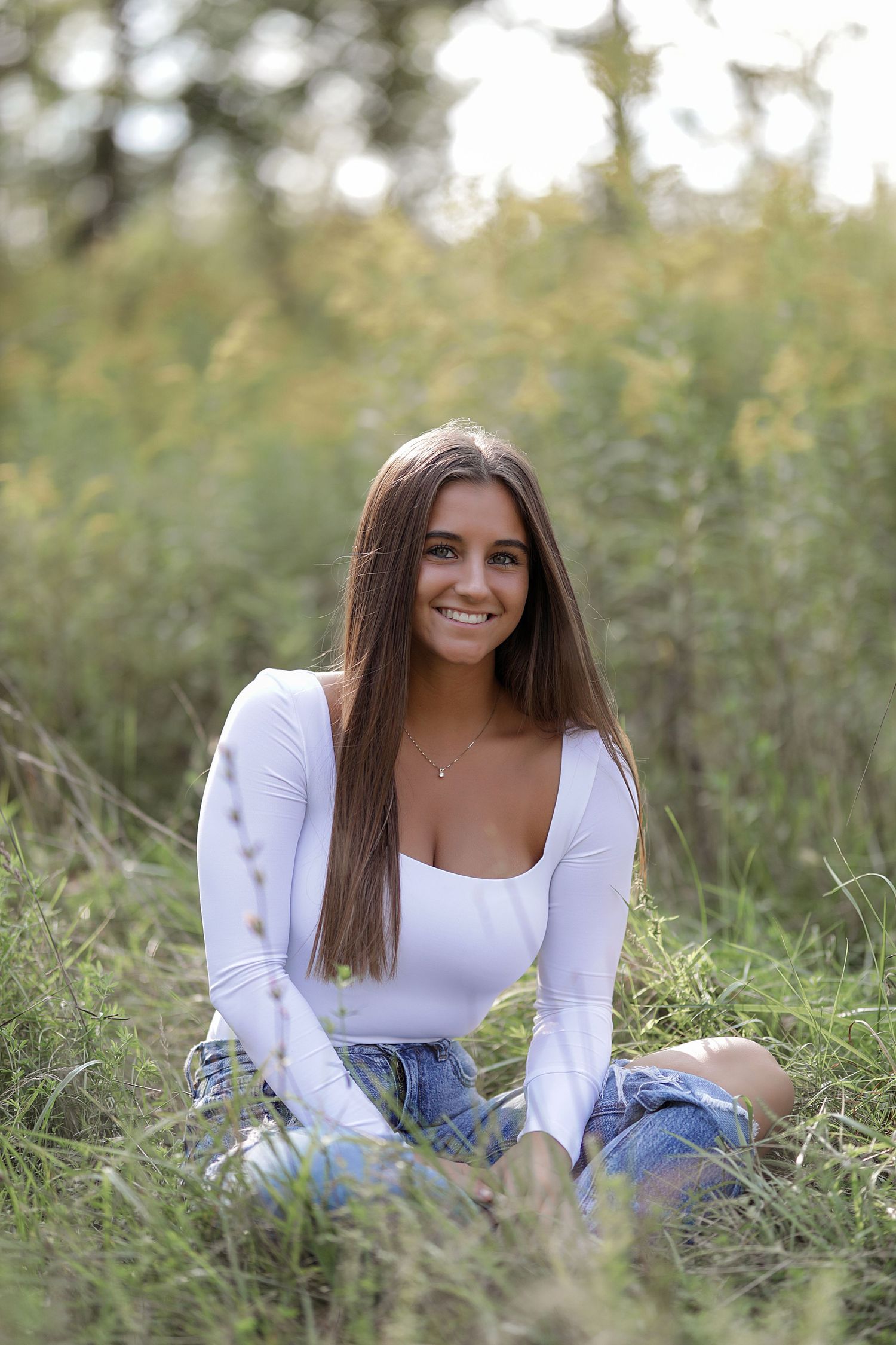 Portrait of someone in a white long sleeve top and blue jeans sitting casually in a grassy outdoor setting.