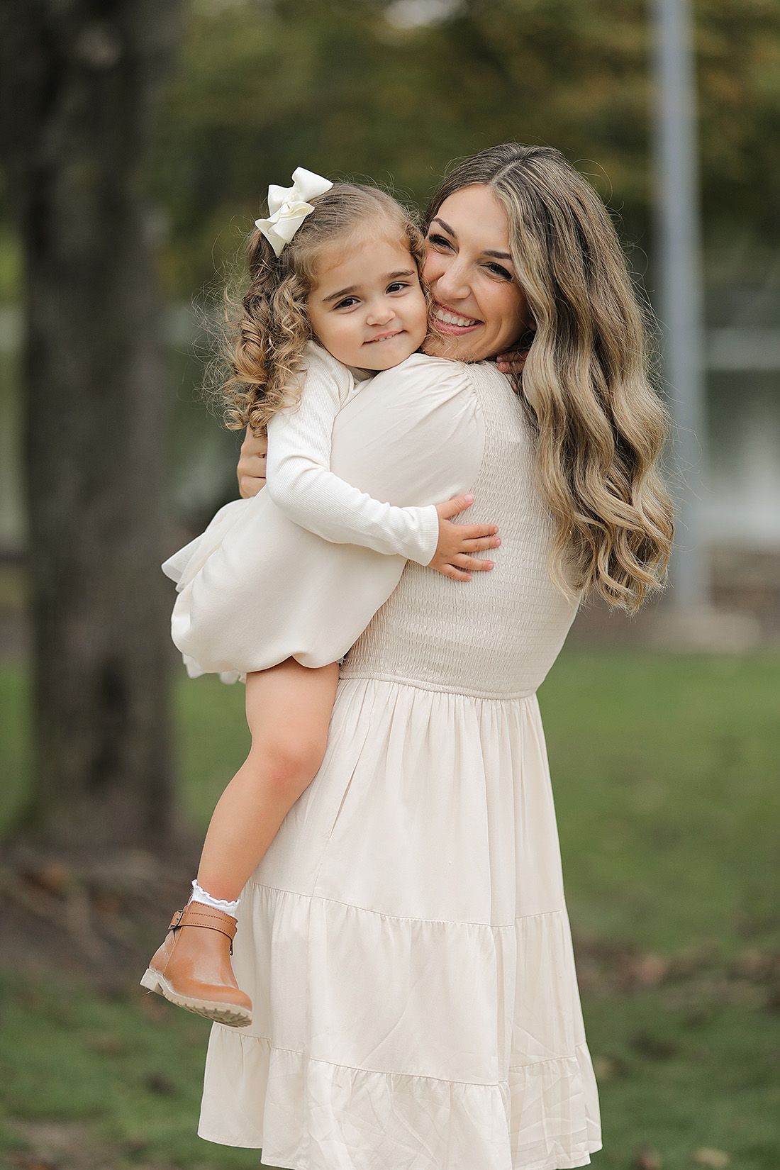 Mother and daughter in matching white dresses pose together for outdoor family portrait.