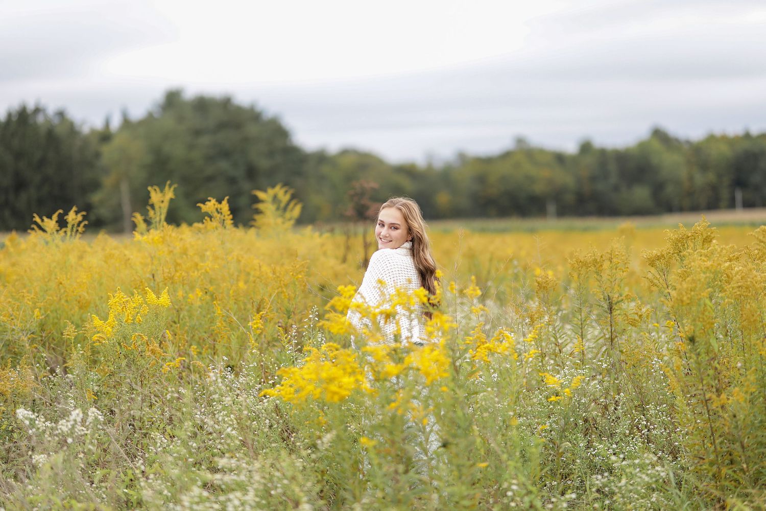 Someone sits in a vibrant yellow wildflower meadow on an overcast day with trees in the background.