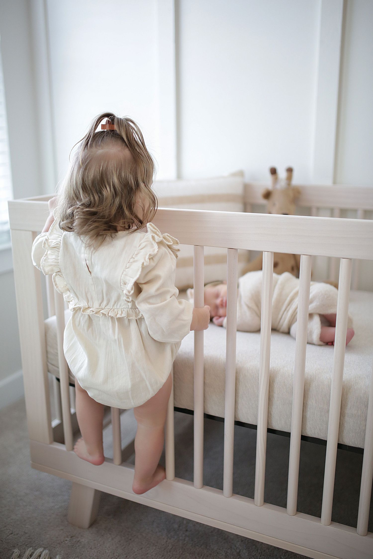 A small child in a cream-colored romper stands at a white crib while looking over the railing.