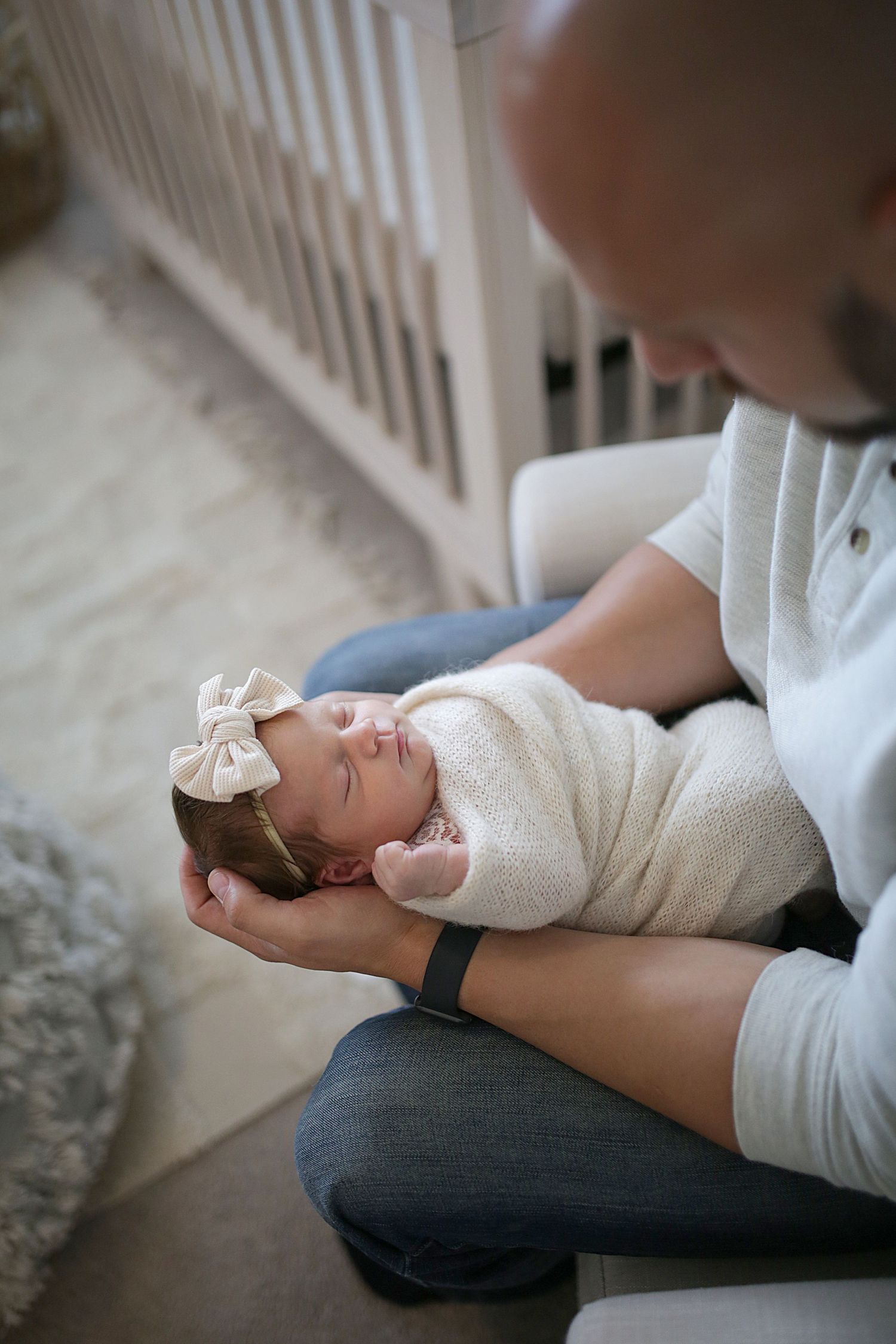 A newborn baby wrapped in white swaddling cloth with a bow headband rests peacefully on an adult's lap.