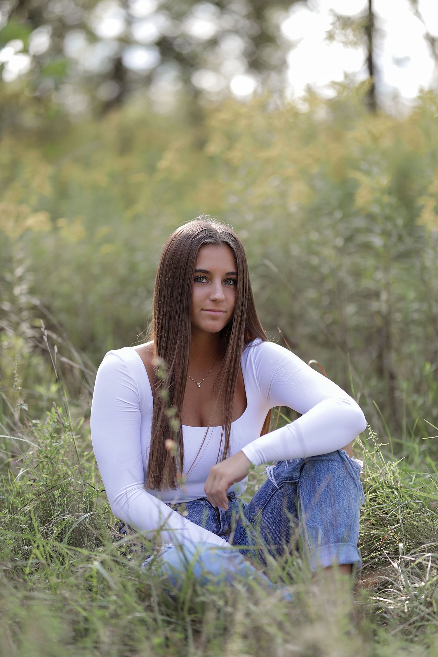A person in a white sweater and jeans sits in a grassy outdoor field during a relaxed portrait session.