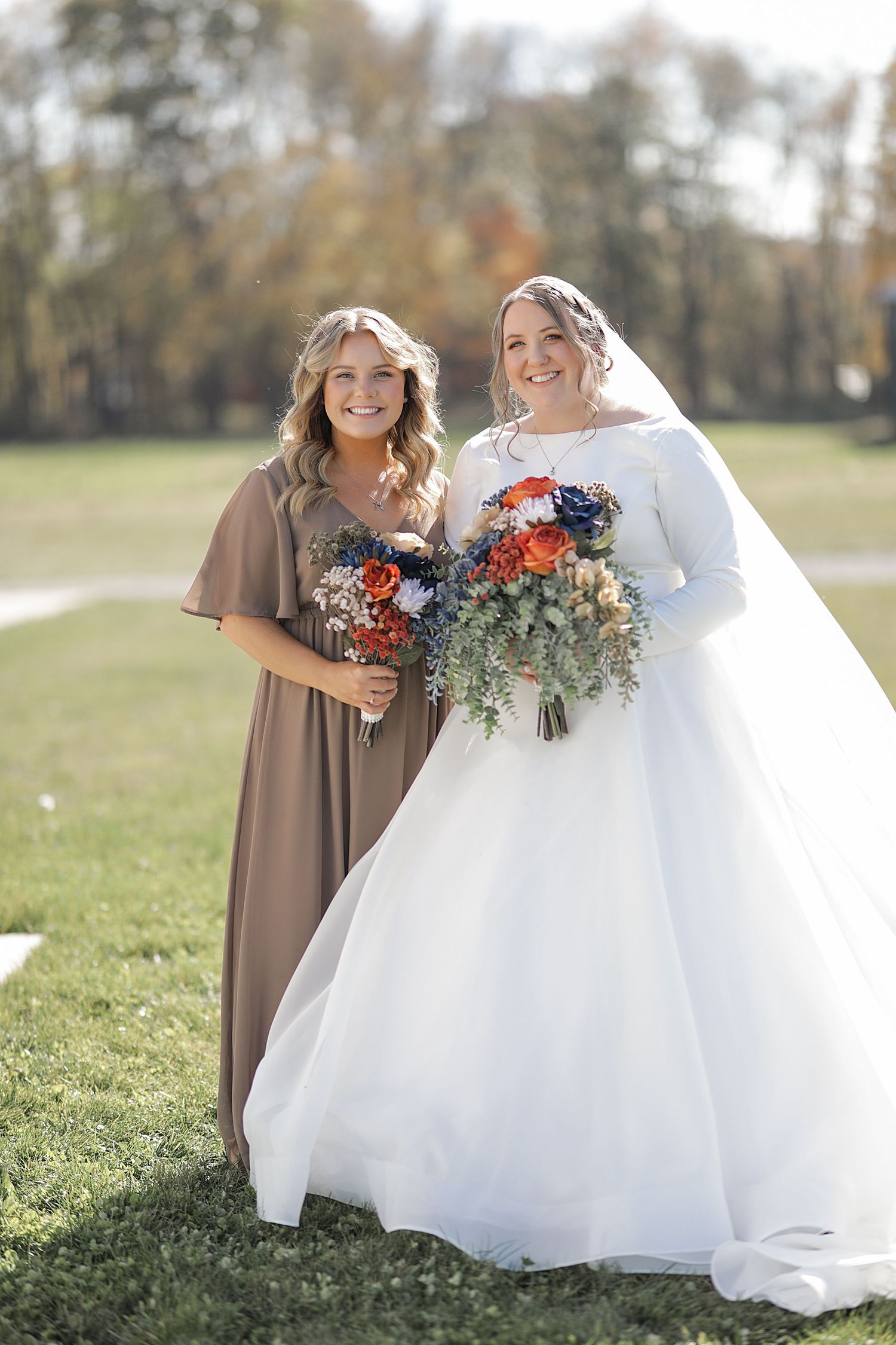 Two wedding party members in brown and white dresses holding fall-colored bouquets outdoors.