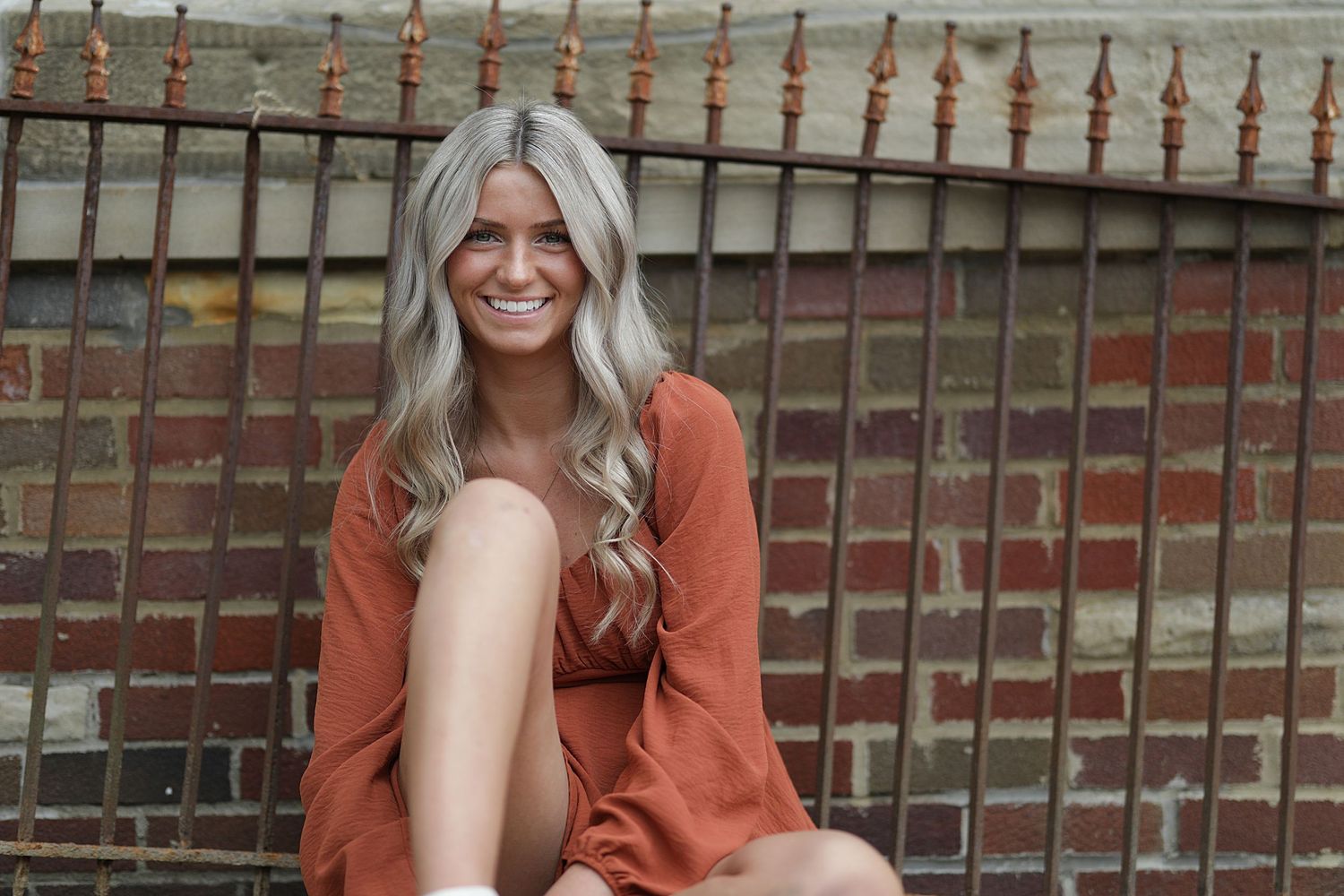 Someone wearing a burnt orange sweater sits against a brick wall and iron fence while smiling warmly.