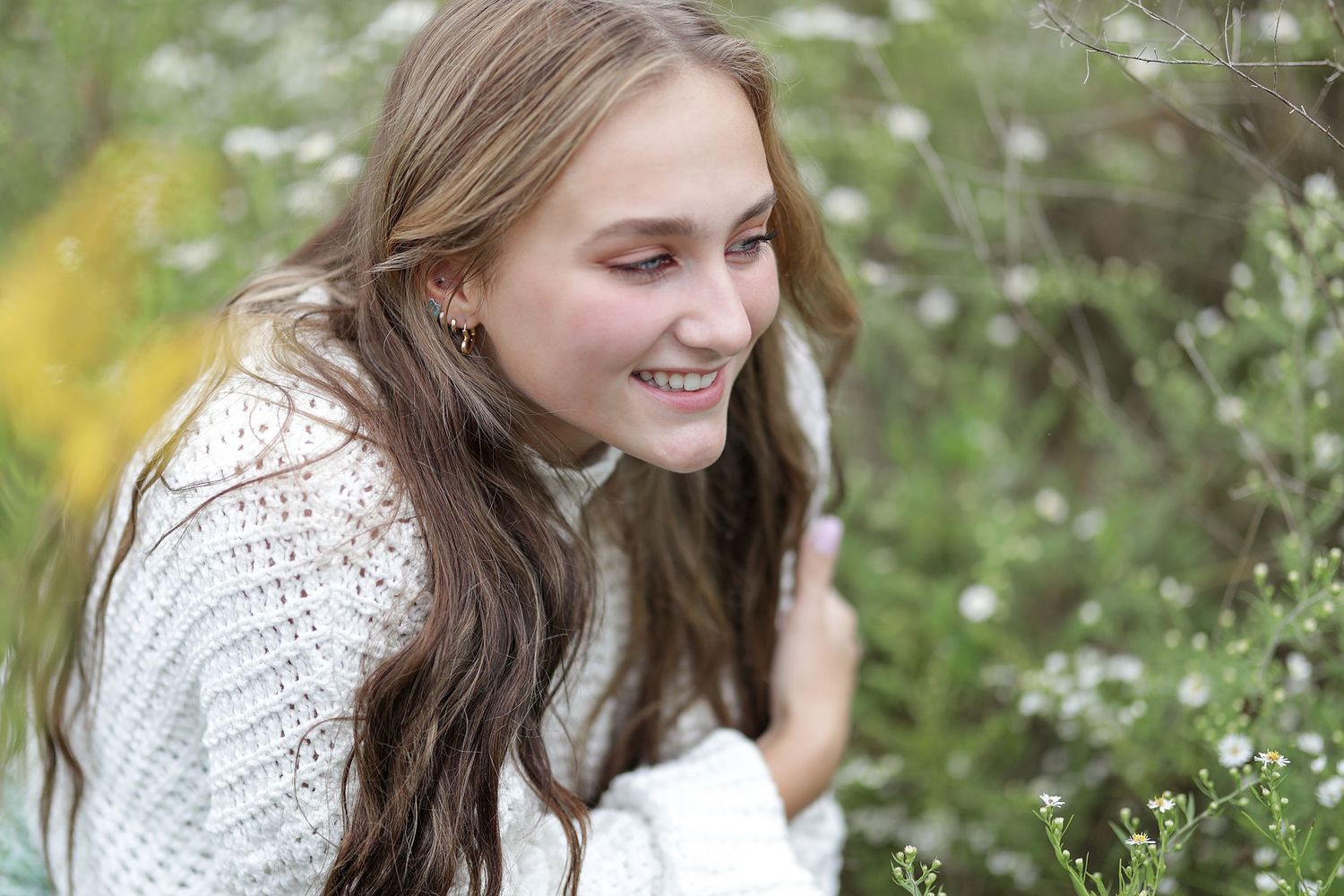 A woman in a white knit sweater smiles while sitting in a meadow with white wildflowers.