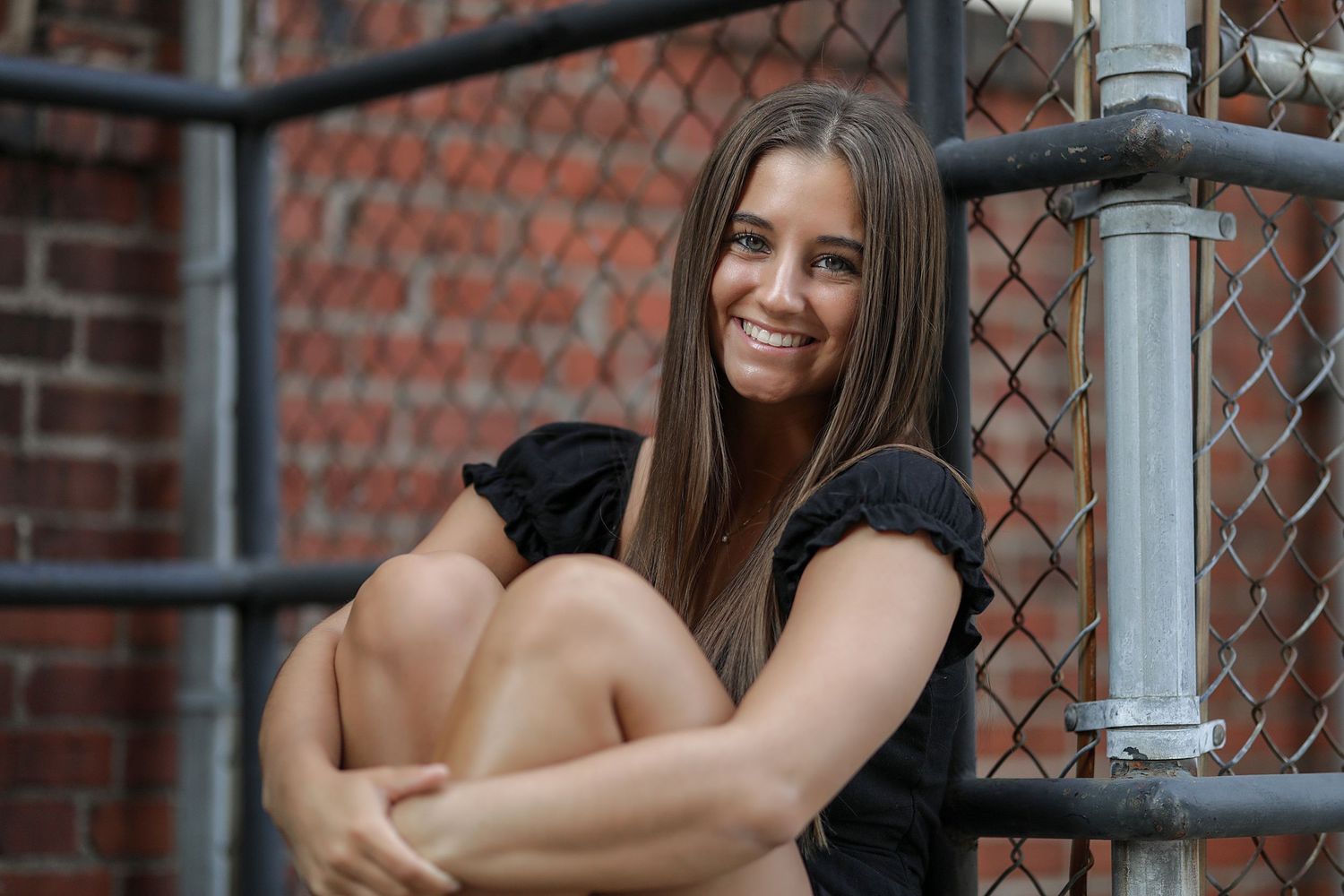Two people laughing together while leaning against a chain link fence with brick wall in background.
