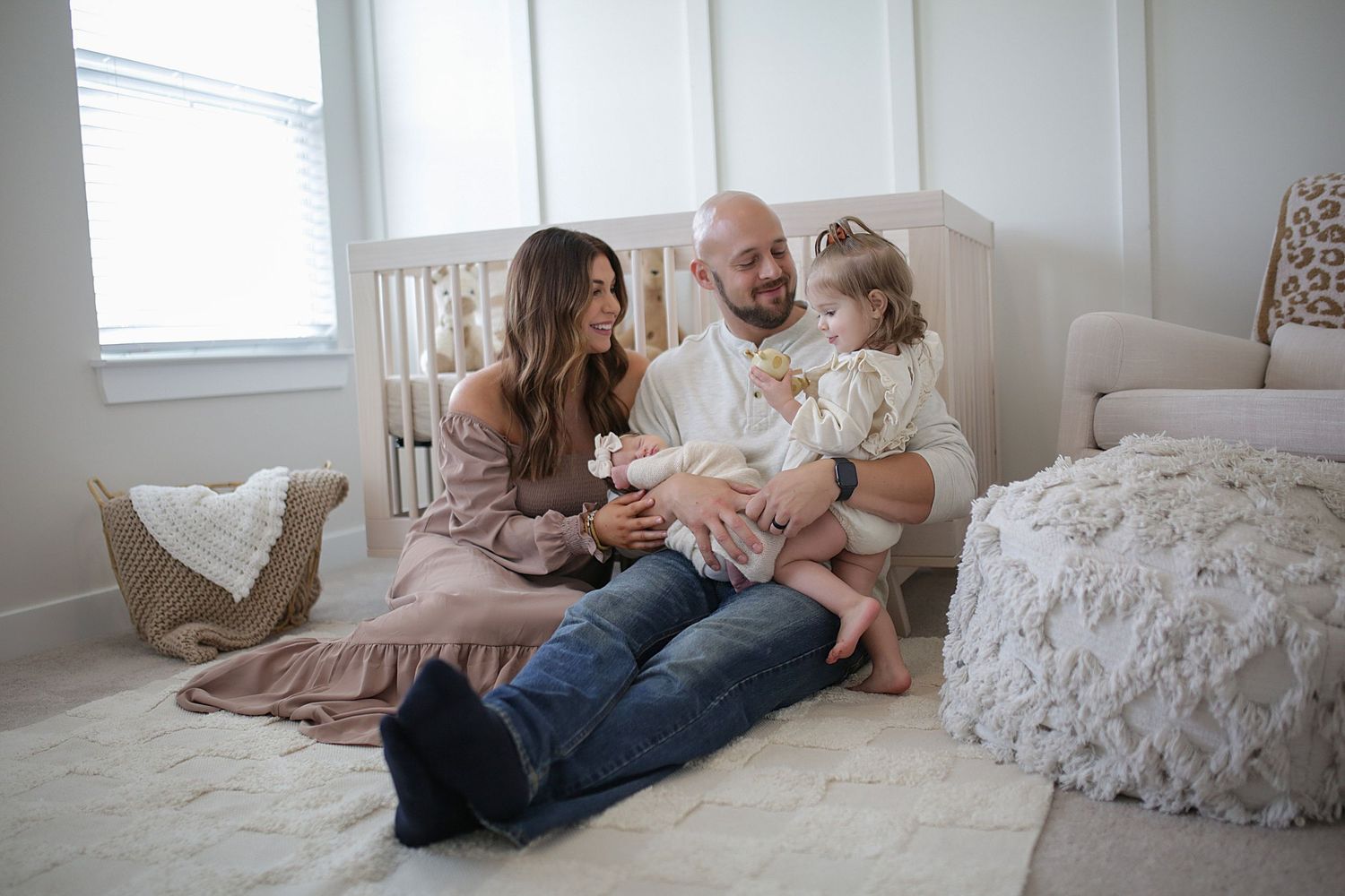 A couple snuggles lovingly with their young child in a cozy sunlit bedroom with white decor.