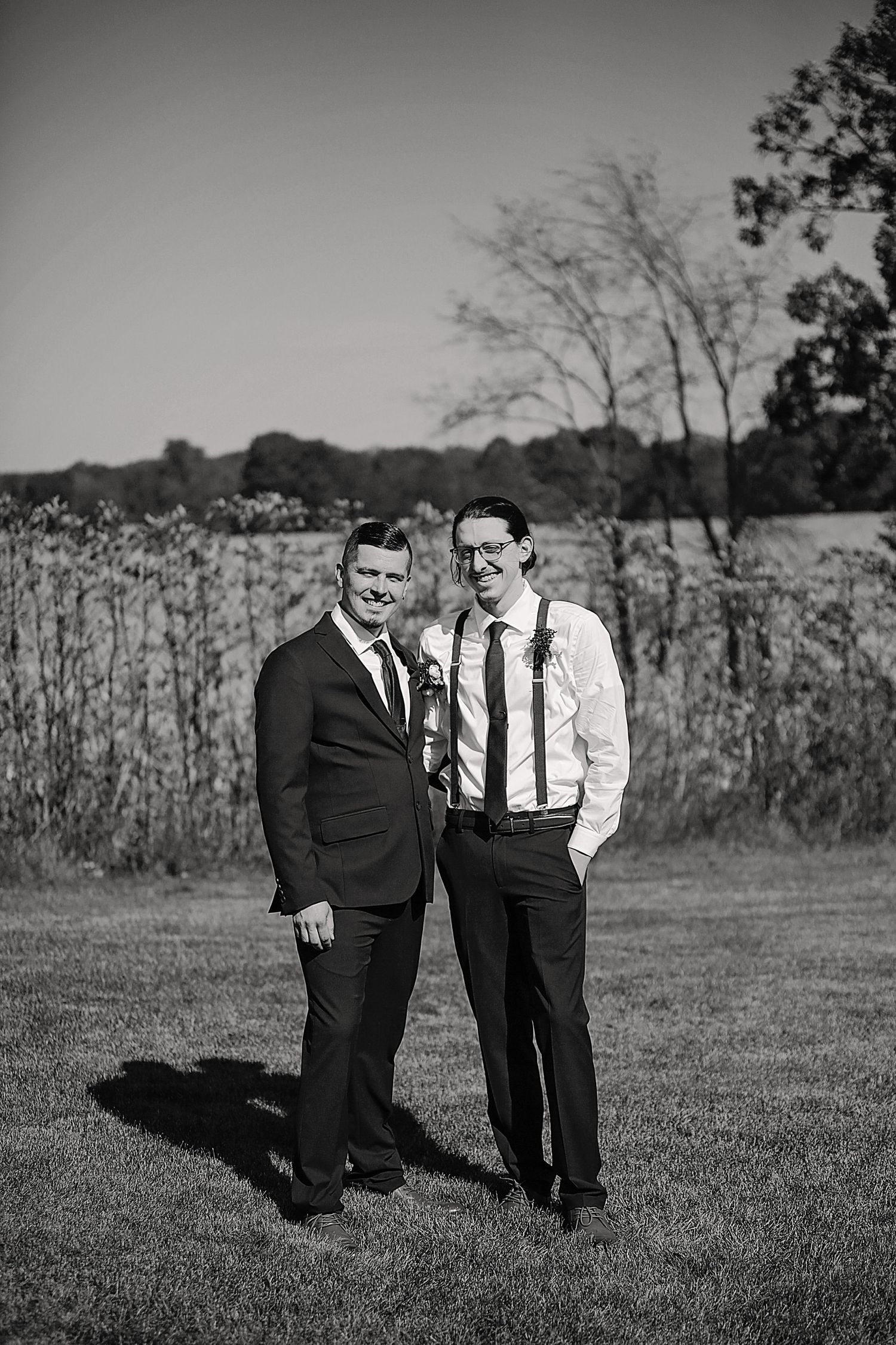 Two groomsmen in formal attire pose together in a black and white outdoor portrait at a wedding.