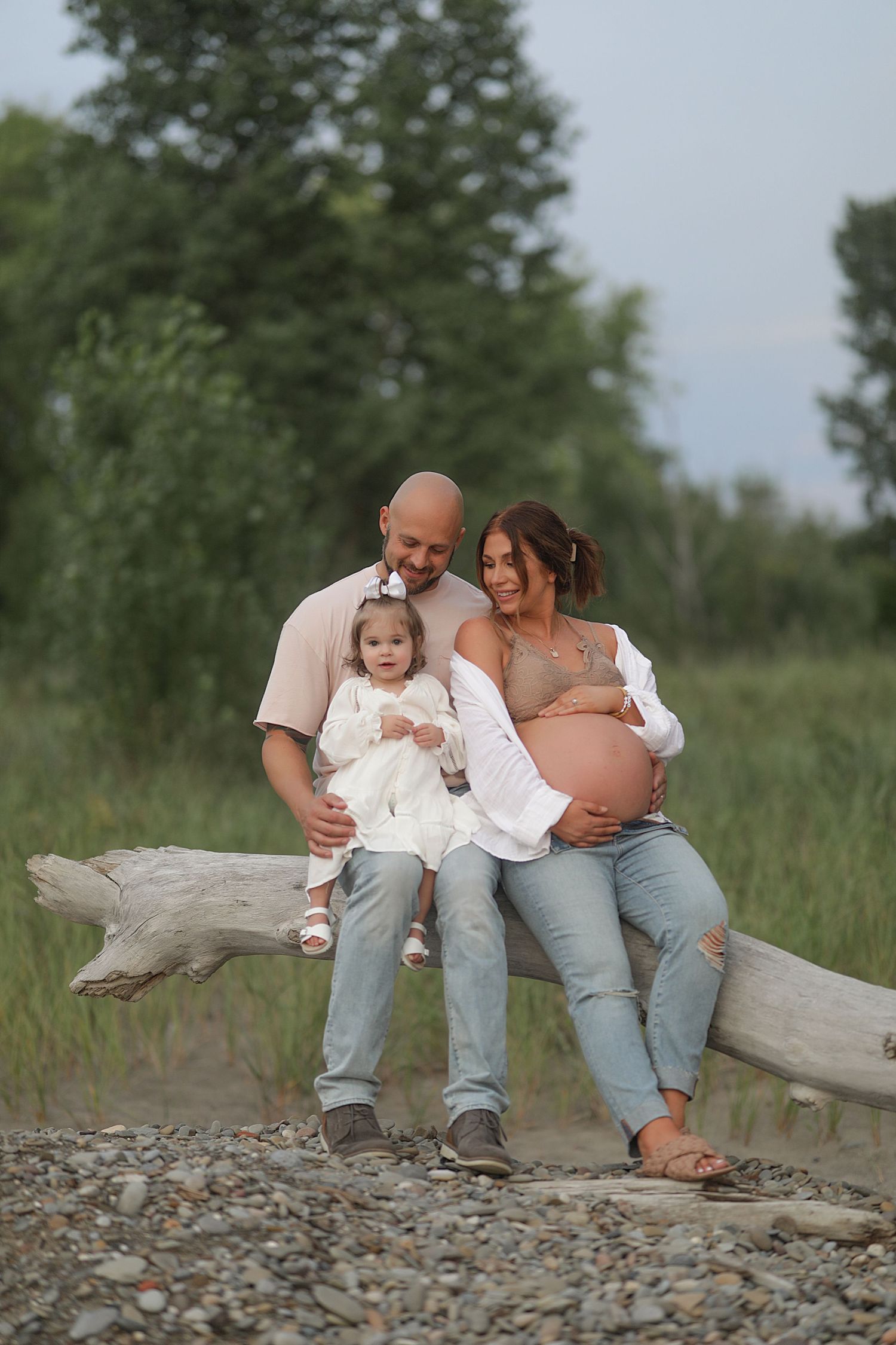 Expecting family sits together on driftwood log at beach during sunset maternity photo session.